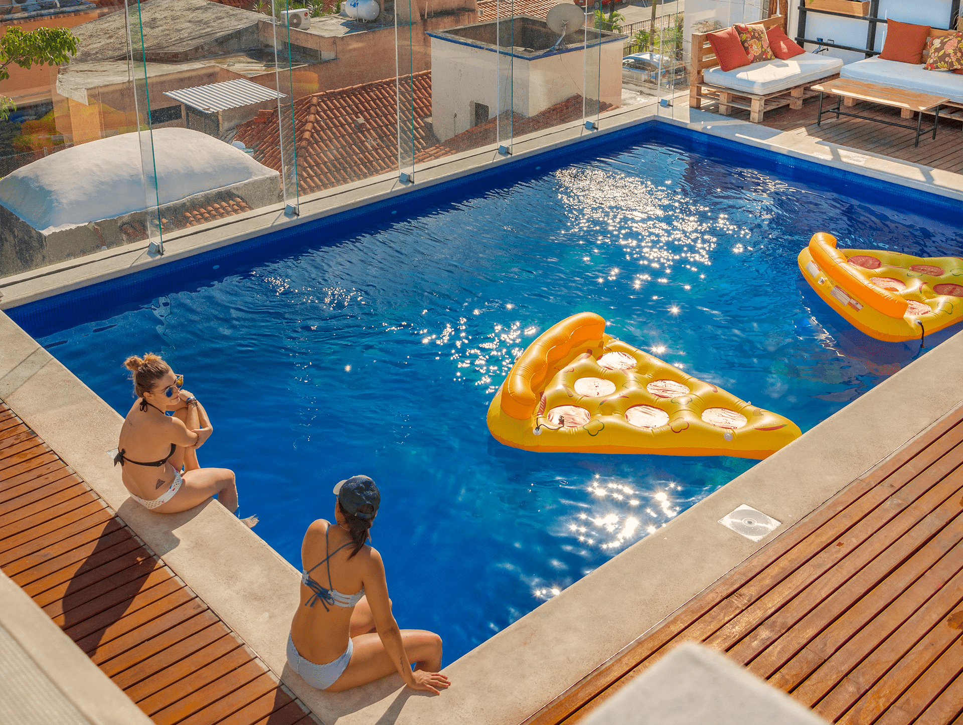 Two women are sitting on the edge of a swimming pool with a pizza float in the water.