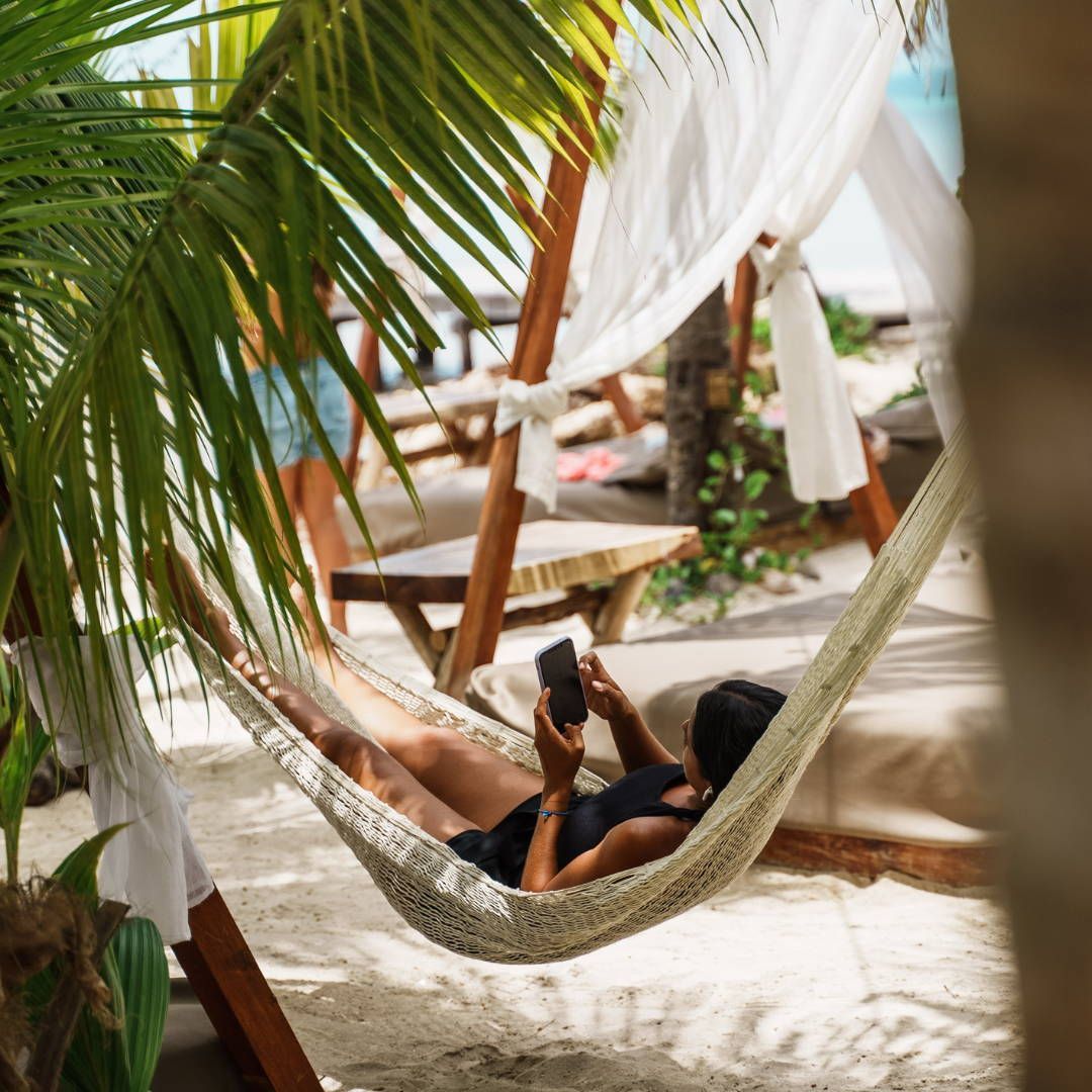 A woman is laying in a hammock on the beach looking at her phone.