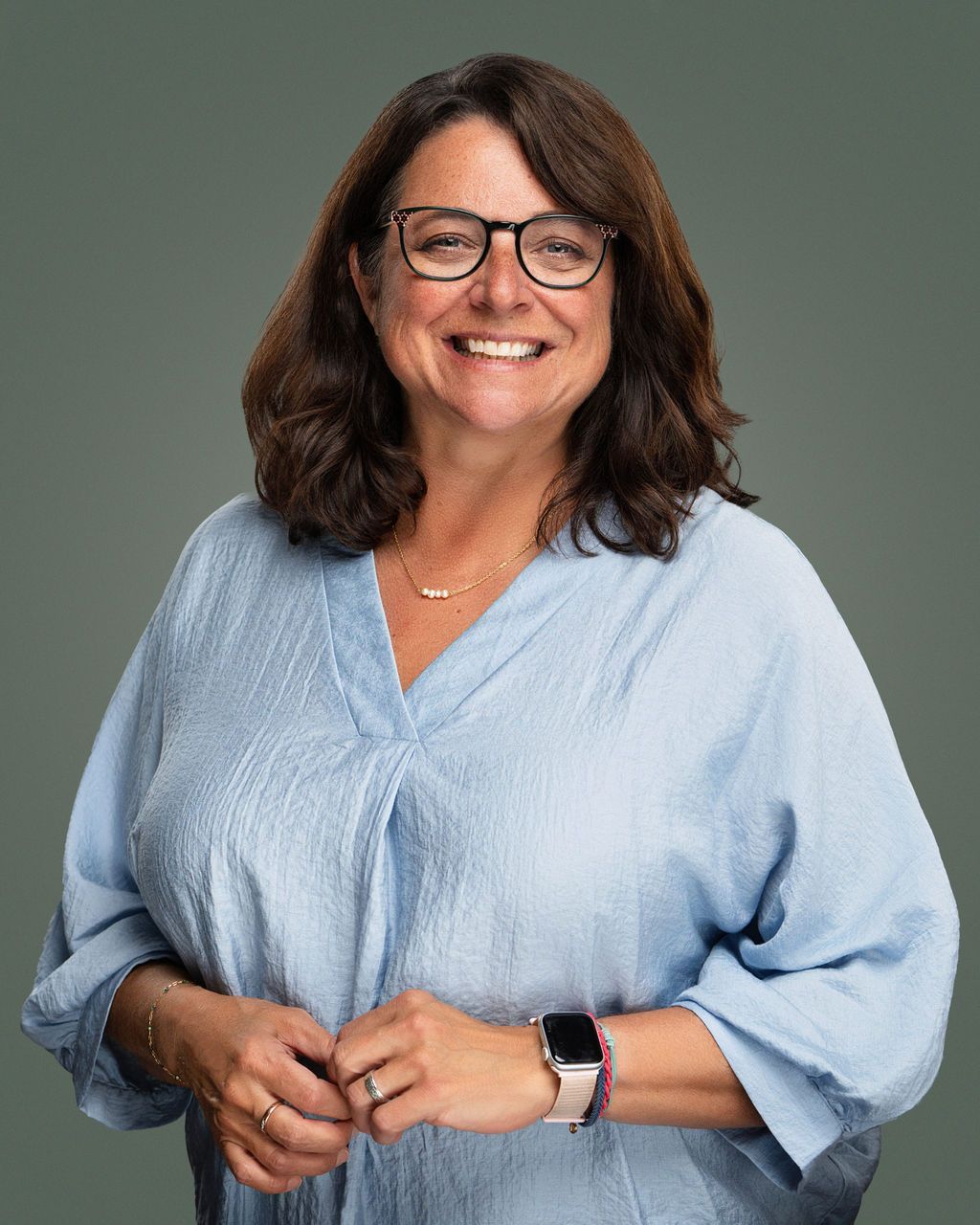Woman with glasses smiles, wearing a light blue top and watch, standing in front of a gray backdrop.