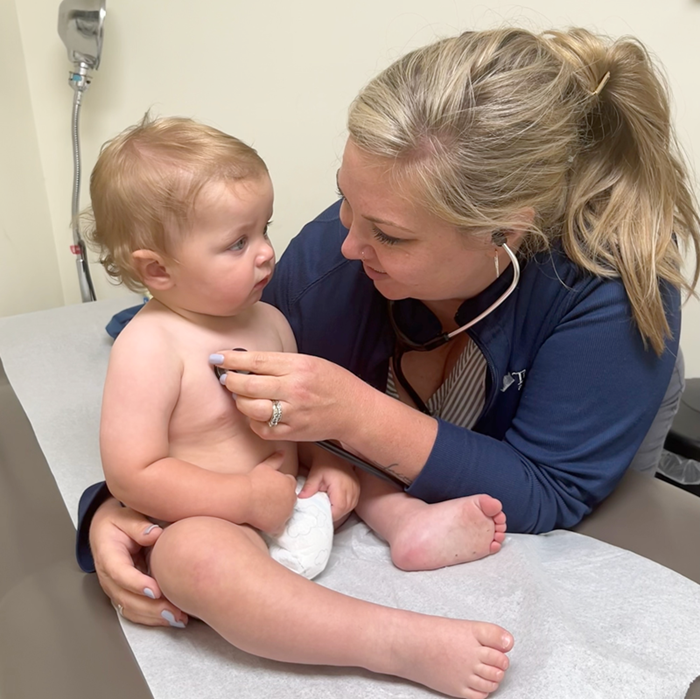 Doctor examining a baby in a clinic exam room, holding a stethoscope to the child’s chest.