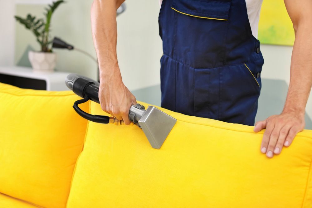 A Man Is Cleaning A Yellow Couch With A Vacuum Cleaner — Encore Carpet Professionals in Parkwood, QLD