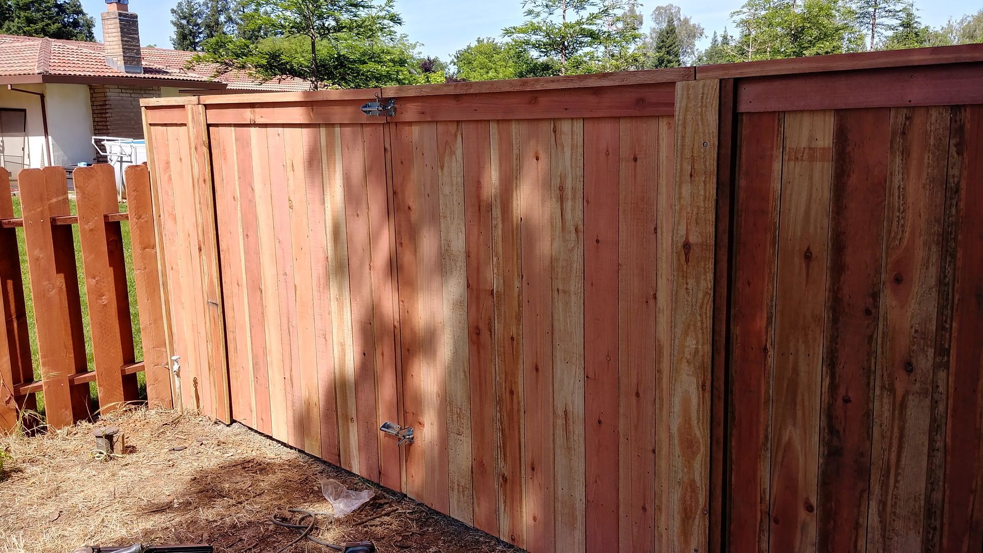 Wooden fence with vertical planks, brown tones. Backed by a gray house and green grass.
