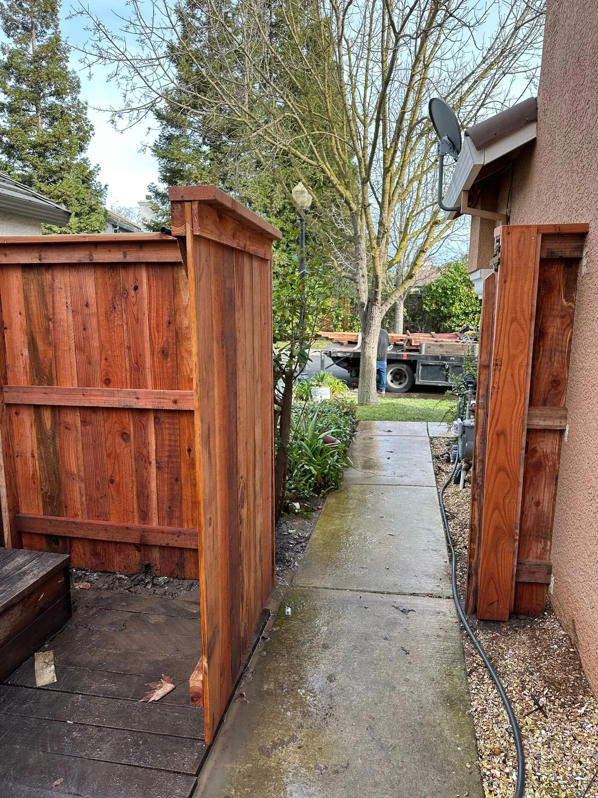 Wooden fence with an open gate next to a house's exterior wall. A concrete walkway leads toward a truck.