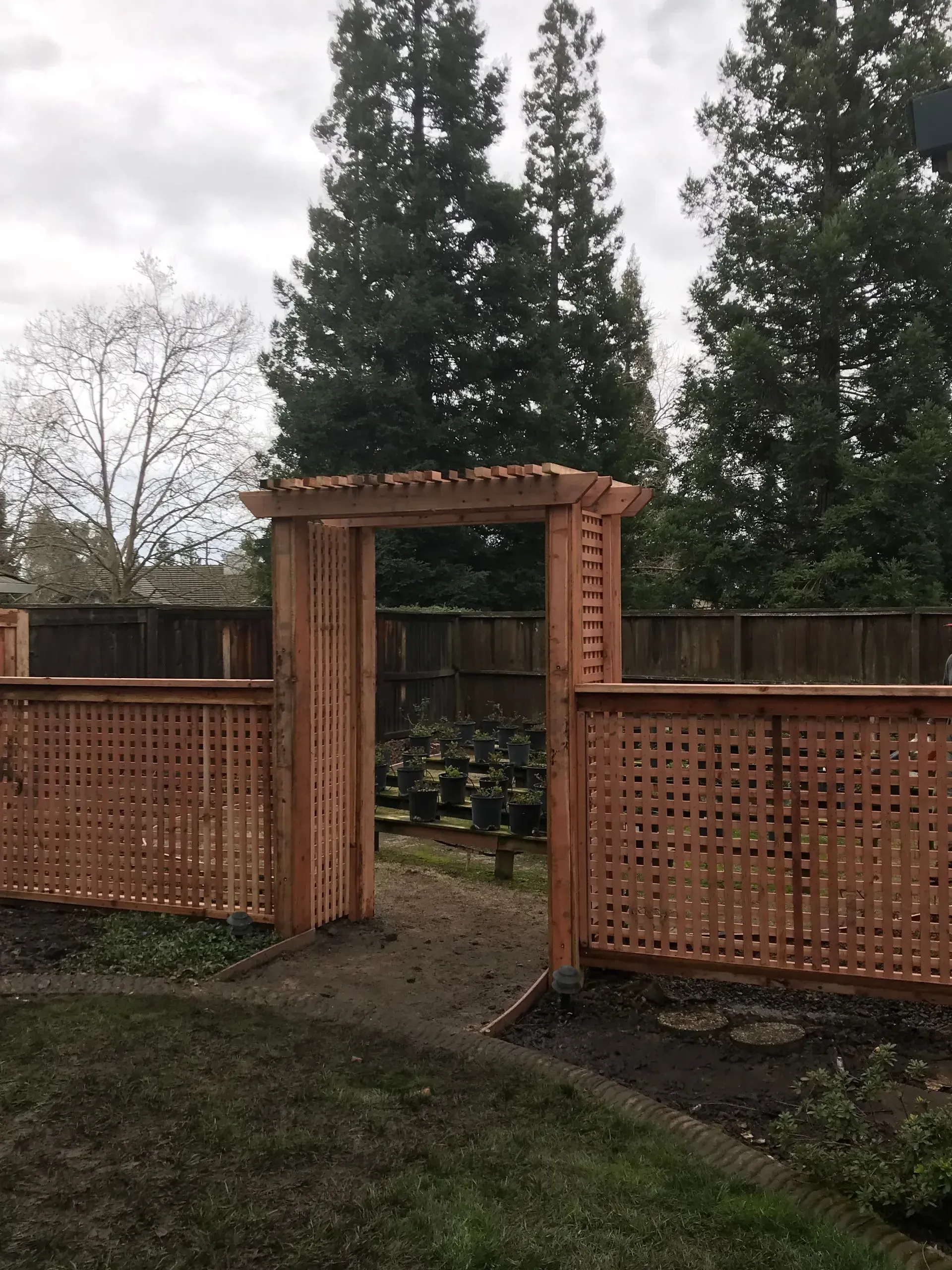 Wooden arbor gateway with lattice-style fence panels, pathway, and trees in background. Cloudy sky.