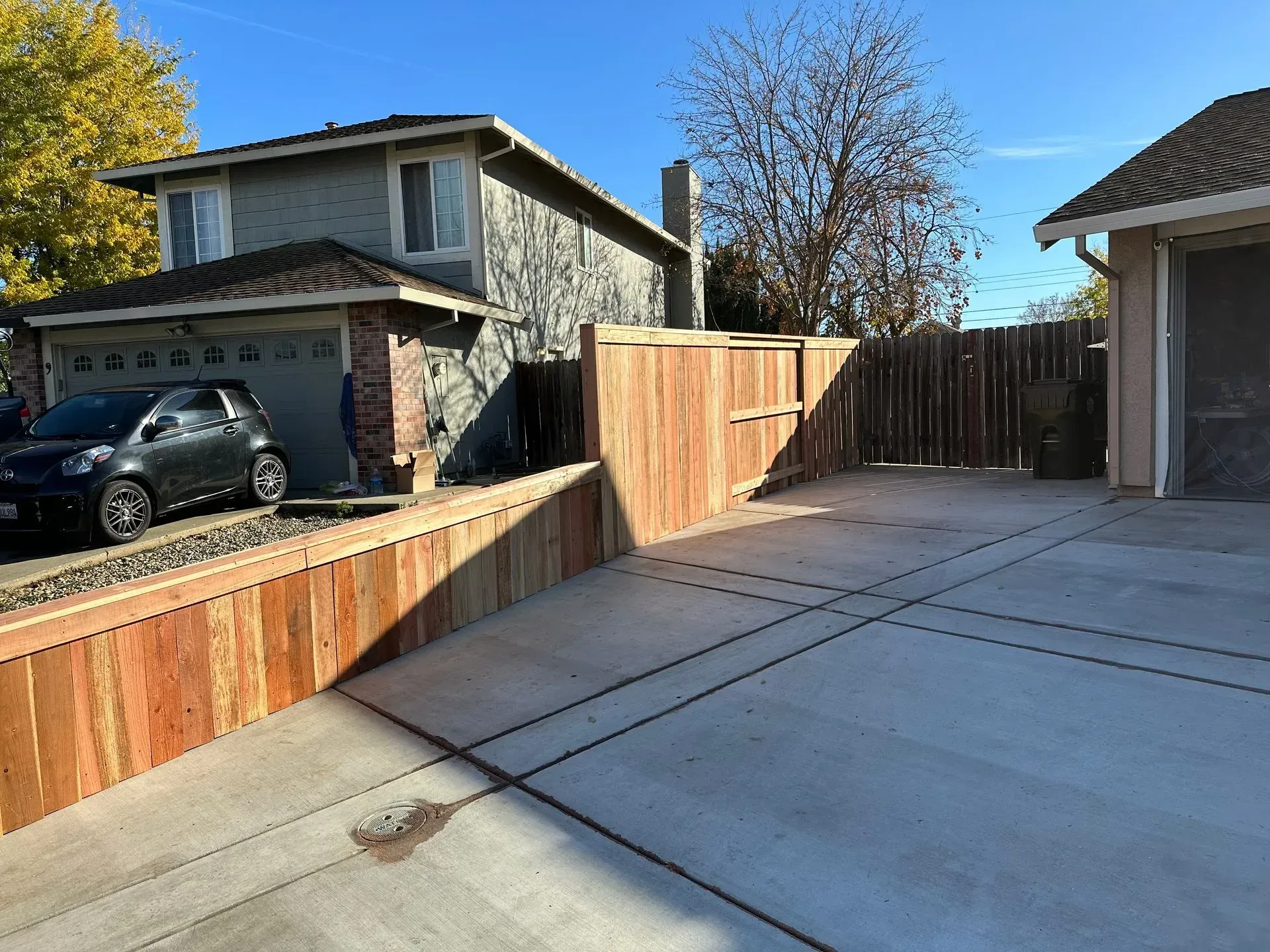 Residential driveway with wooden retaining wall and fence, cars, and a house. Concrete pavement.