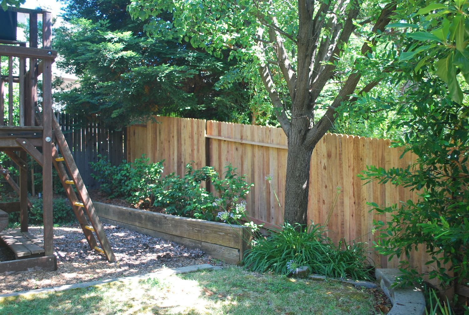 Wooden gate and fence, stained reddish-brown, in a yard with concrete and greenery.