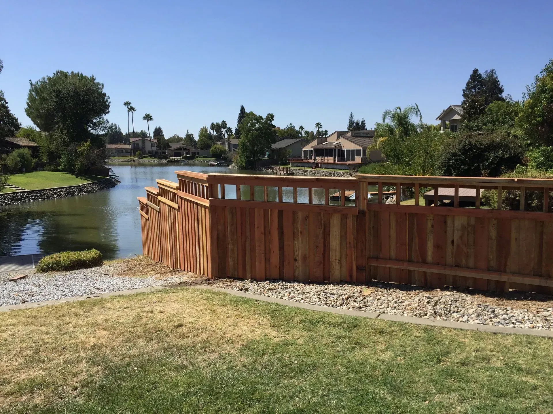 Wooden fence along a lake, with houses visible in the background under a blue sky.