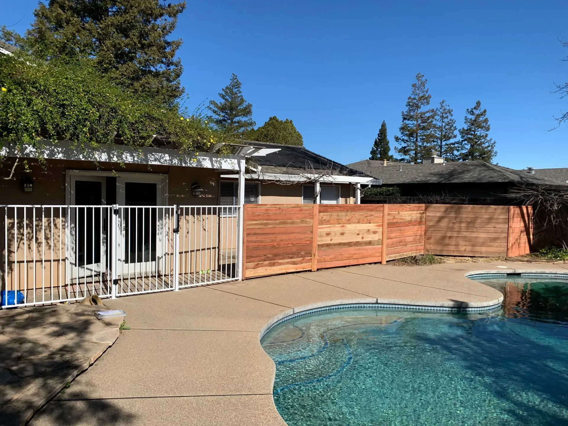 Backyard with pool, patio, wooden fence, and house under a blue sky.