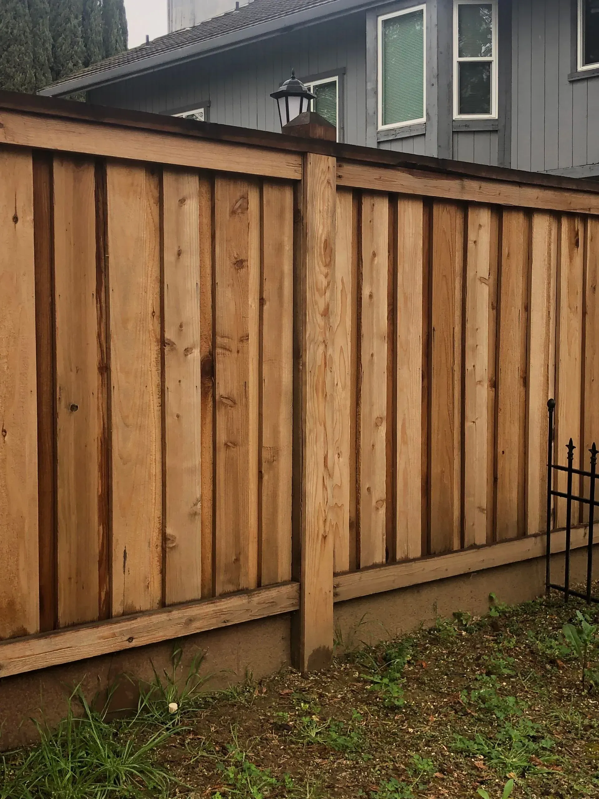 Wooden fence with vertical planks, brown tones. Backed by a gray house and green grass.