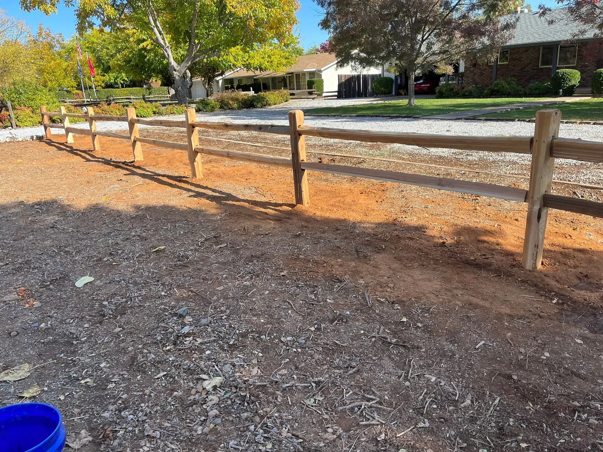 Wooden split-rail fence along a dirt area, with a residential street and houses in the background.