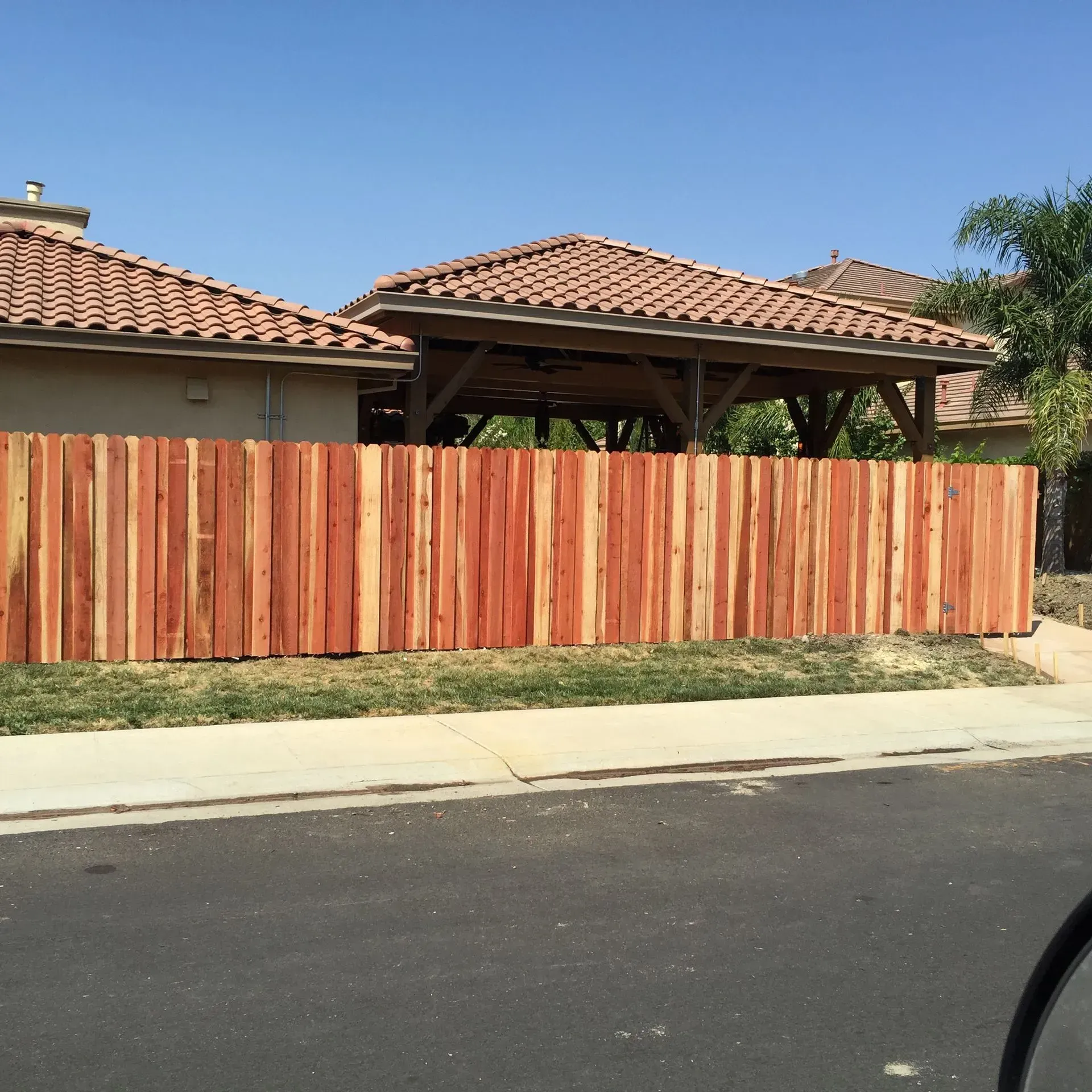 Red wooden privacy fence in front of a house with a gazebo and tile roof on a sunny day.