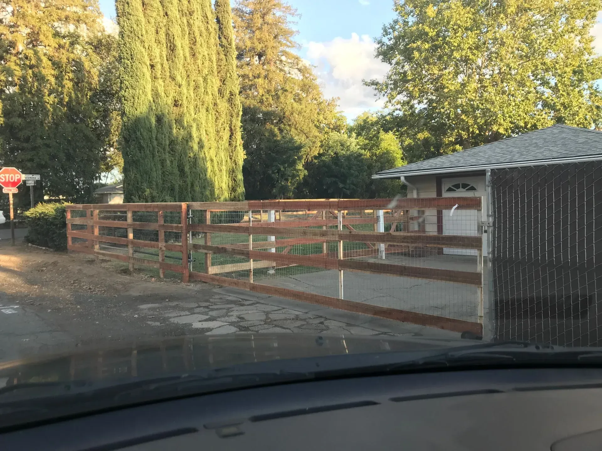 Wooden fence and gate in front of a house, with trees in the background, viewed from inside a vehicle.