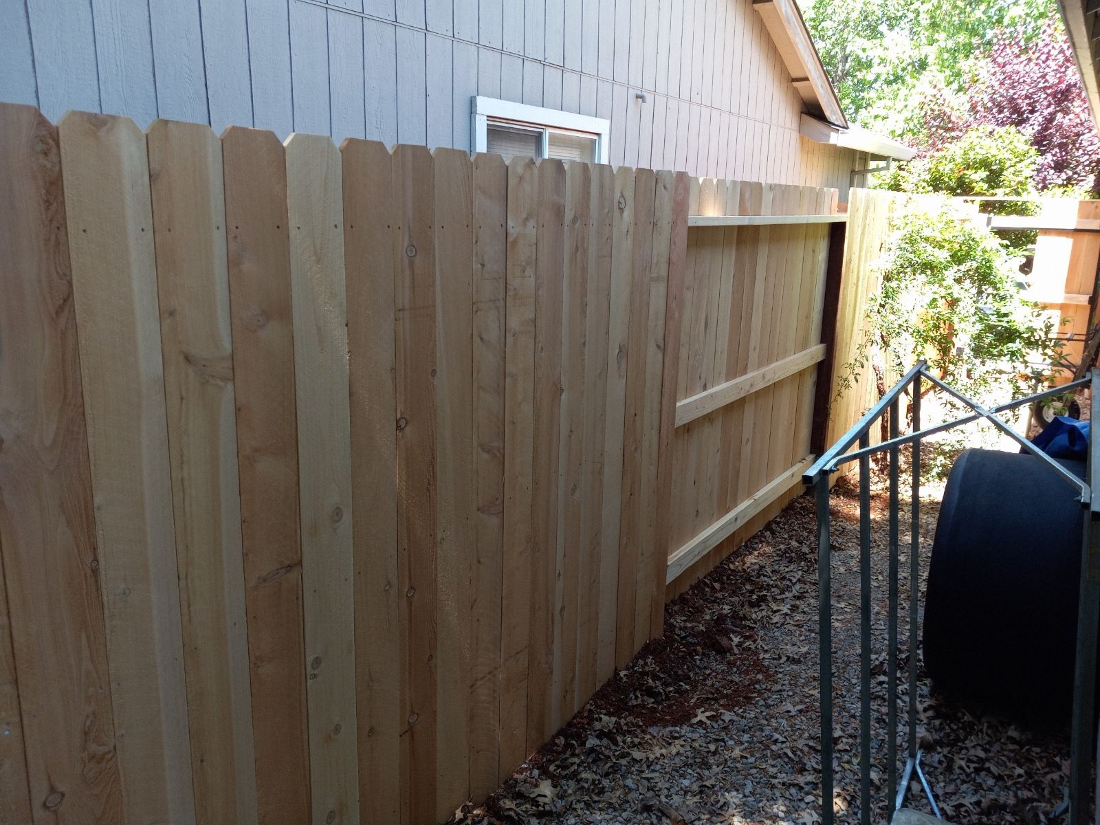 Residential street with a wooden fence, trees, and a house under a blue sky.