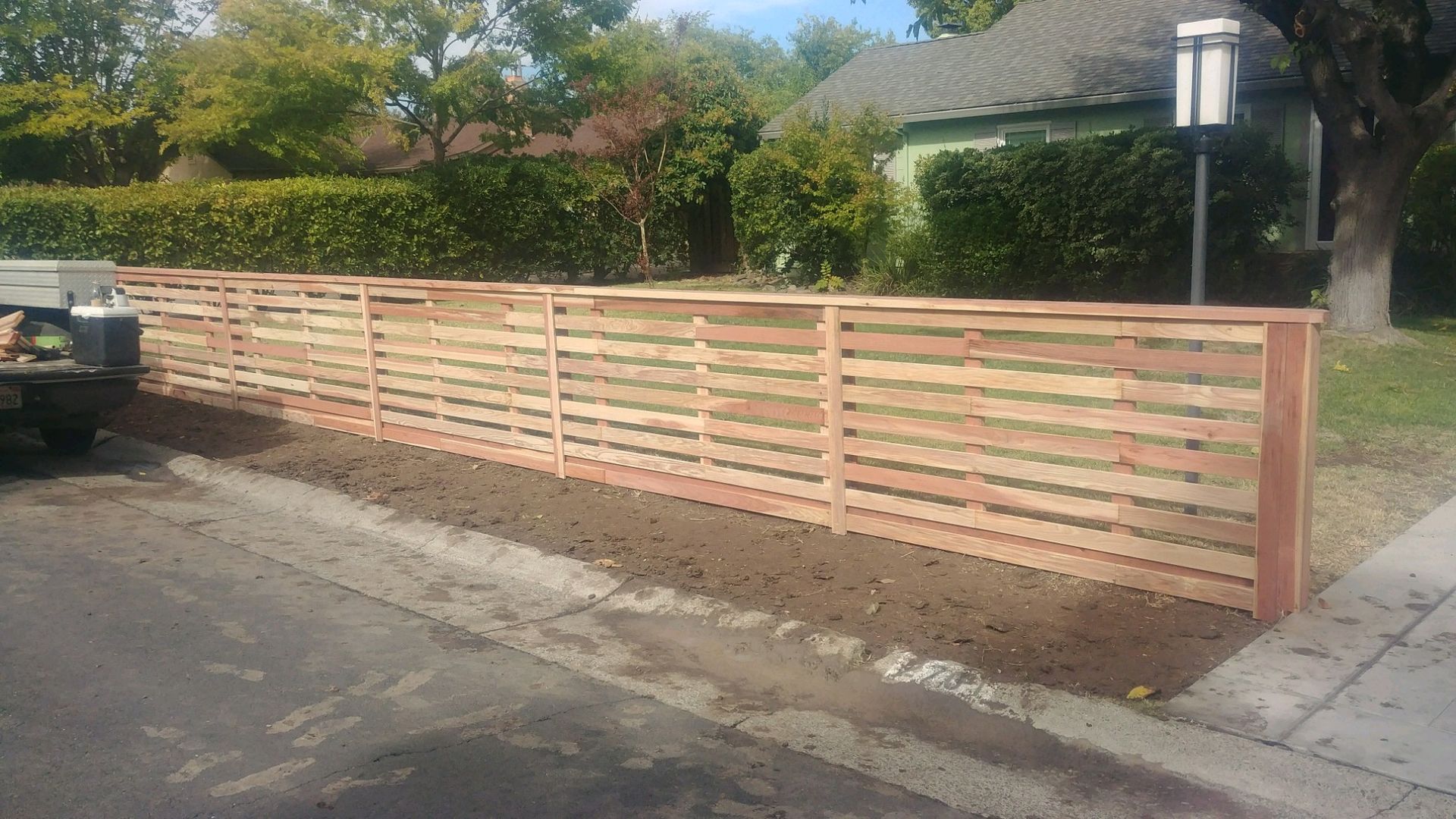 Residential street with a wooden fence, trees, and a house under a blue sky.