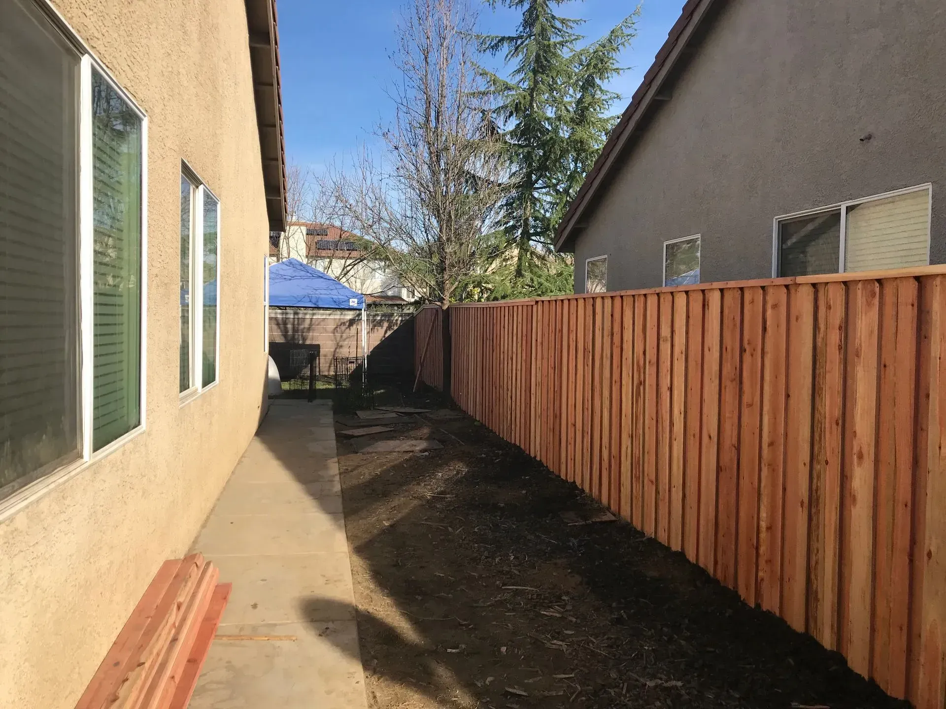 Backyard with a brown wooden fence, a tan house, a bare dirt strip, and a sunny sky.