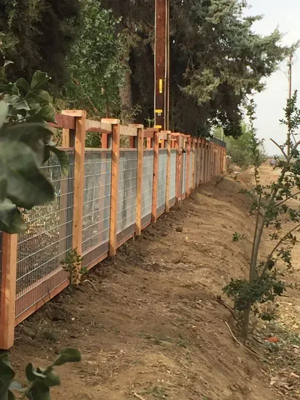 Wooden fence with wire mesh panels, next to a dirt embankment, under trees.