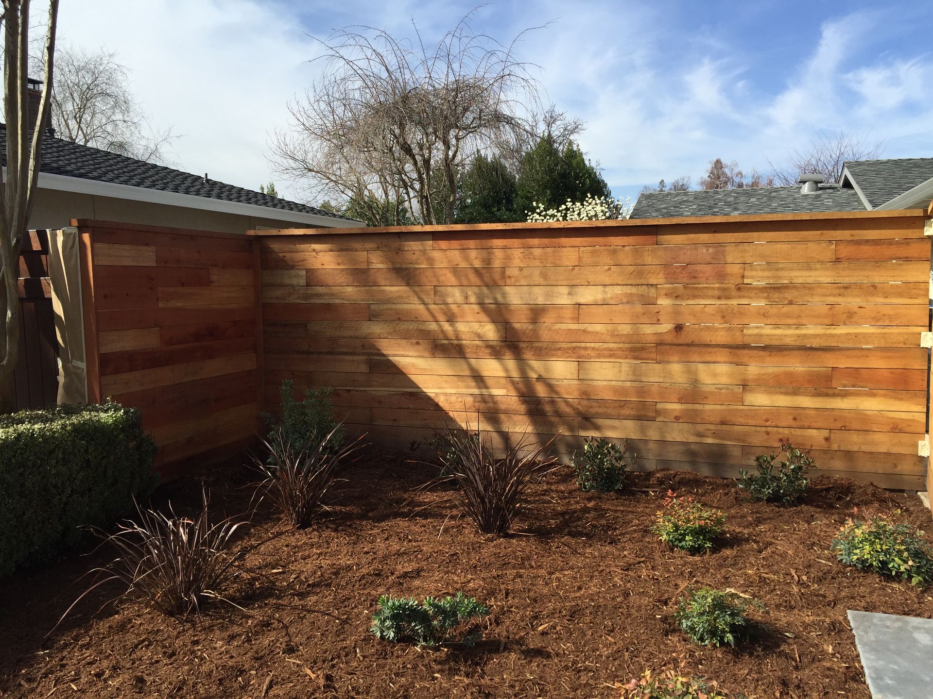 Wooden fence in a yard with mulch and small plants, under a blue sky.