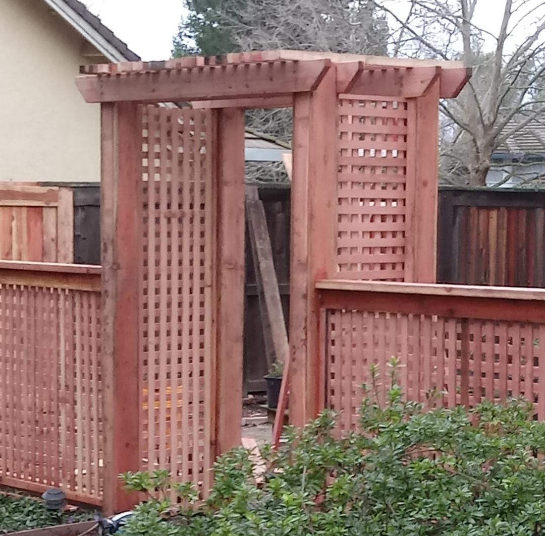Red wooden arbor gate with lattice panels, surrounded by a matching fence, in an outdoor setting.