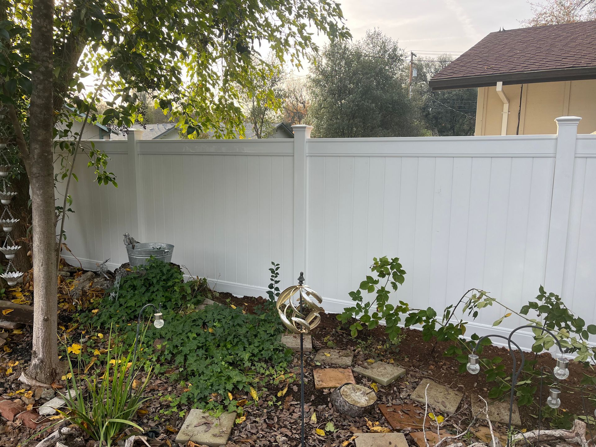 Backyard with a brown wooden fence, a tan house, a bare dirt strip, and a sunny sky.