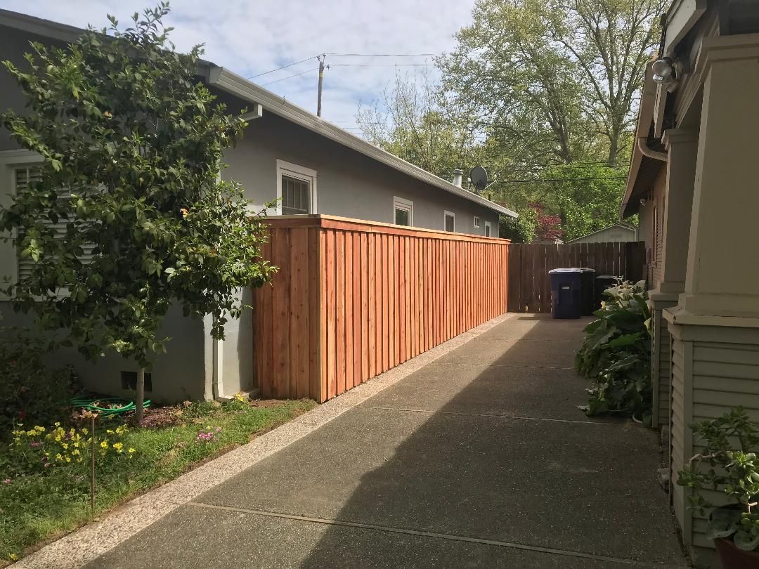 Wooden gate and fence, stained reddish-brown. Gate has a diagonal brace. A concrete slab and dirt are in the foreground.