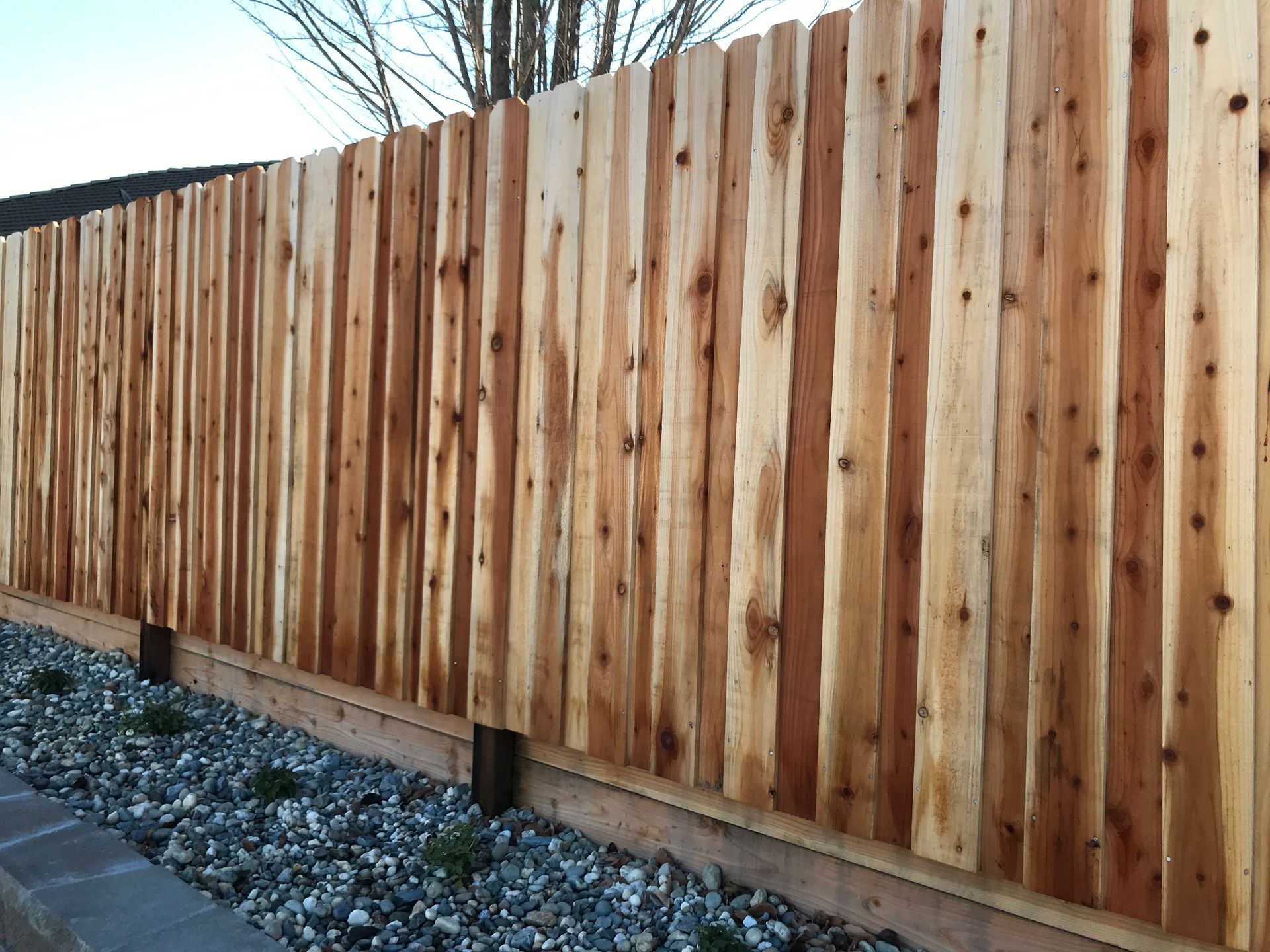 A narrow driveway with a new wooden fence, a gray house, and a few trees on a sunny day.