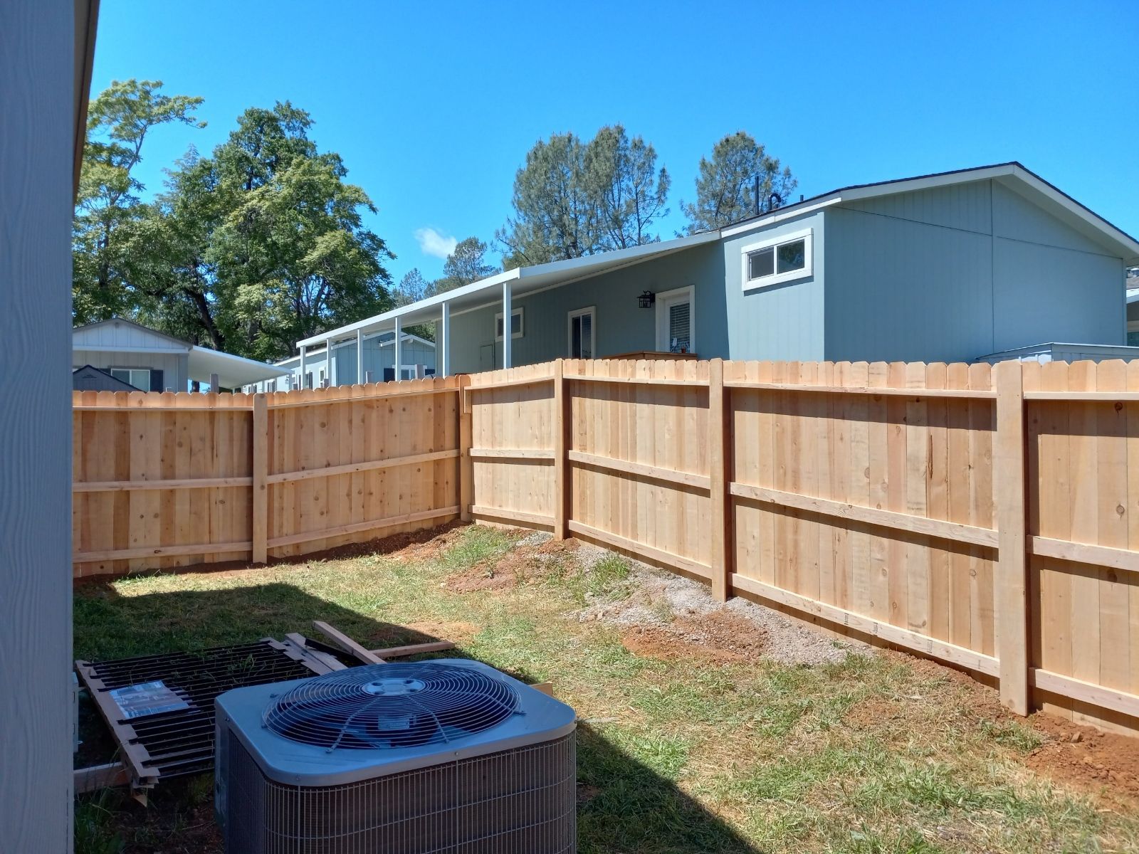 A narrow driveway with a new wooden fence, a gray house, and a few trees on a sunny day.