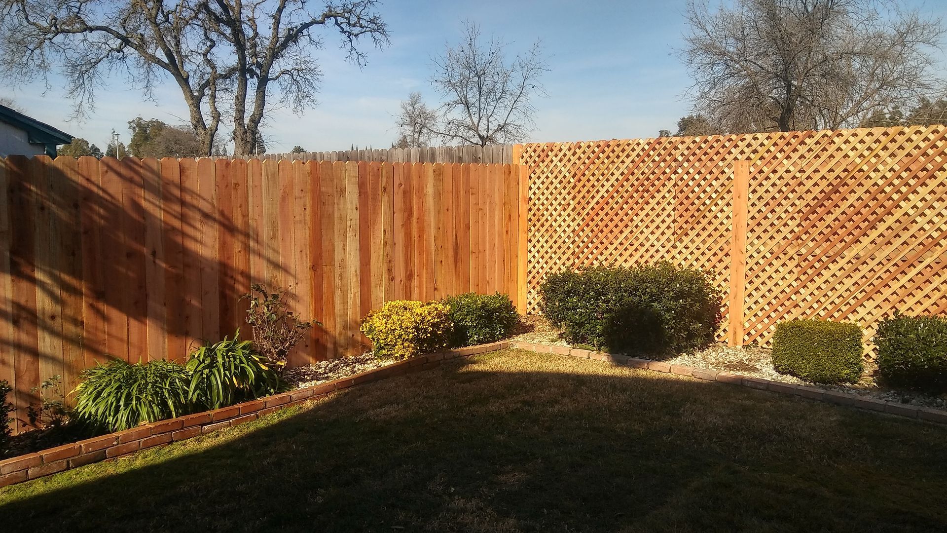 Wooden fence with vertical planks, brown tones. Backed by a gray house and green grass.