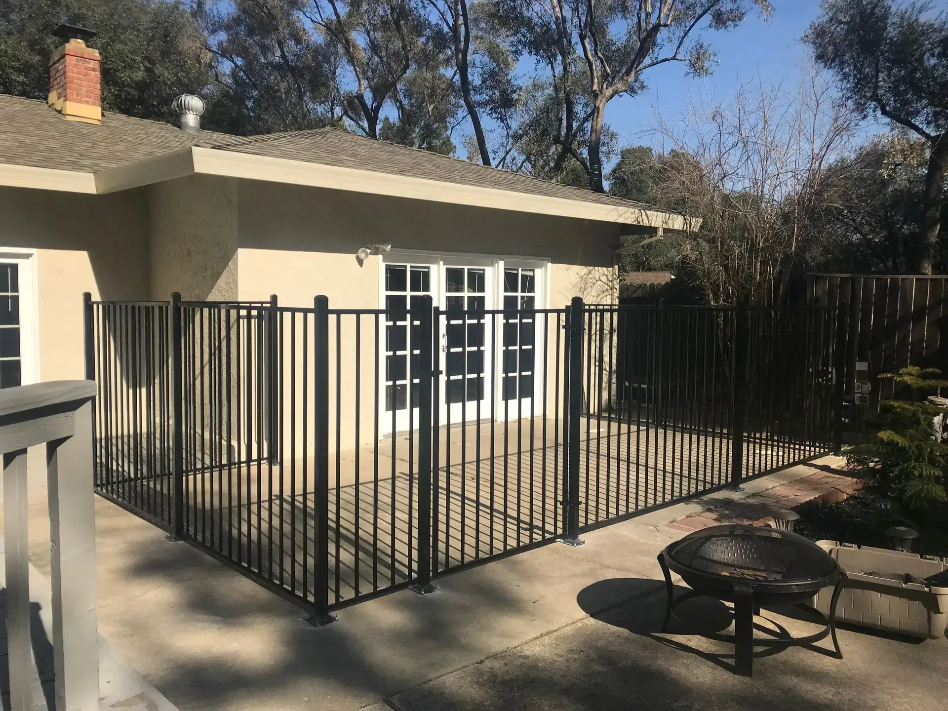 Black metal fence encloses a patio with white French doors. Fire pit and beige house visible.