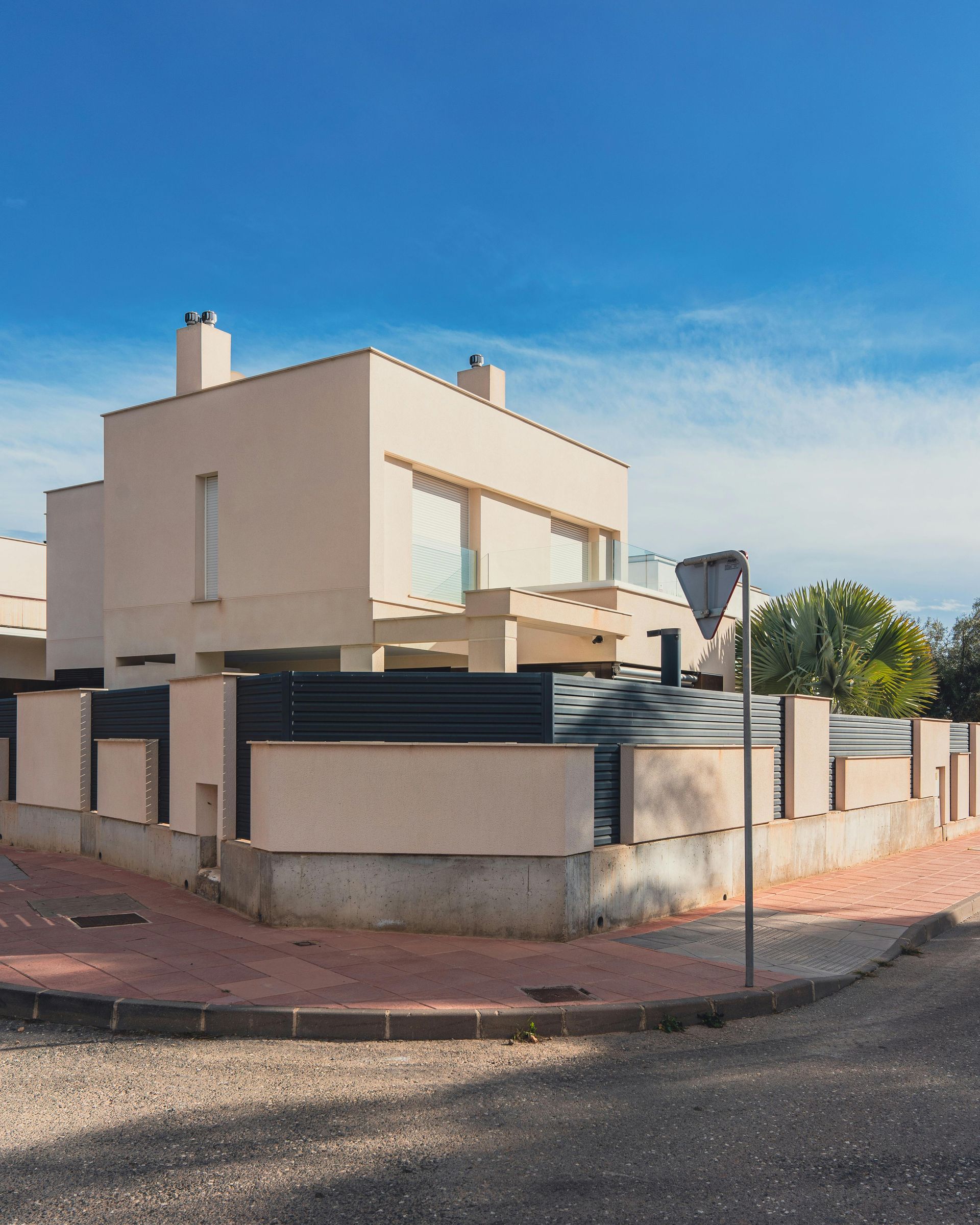 A modern, beige, multi-level house with dark horizontal fencing and chimneys under a bright blue sky.