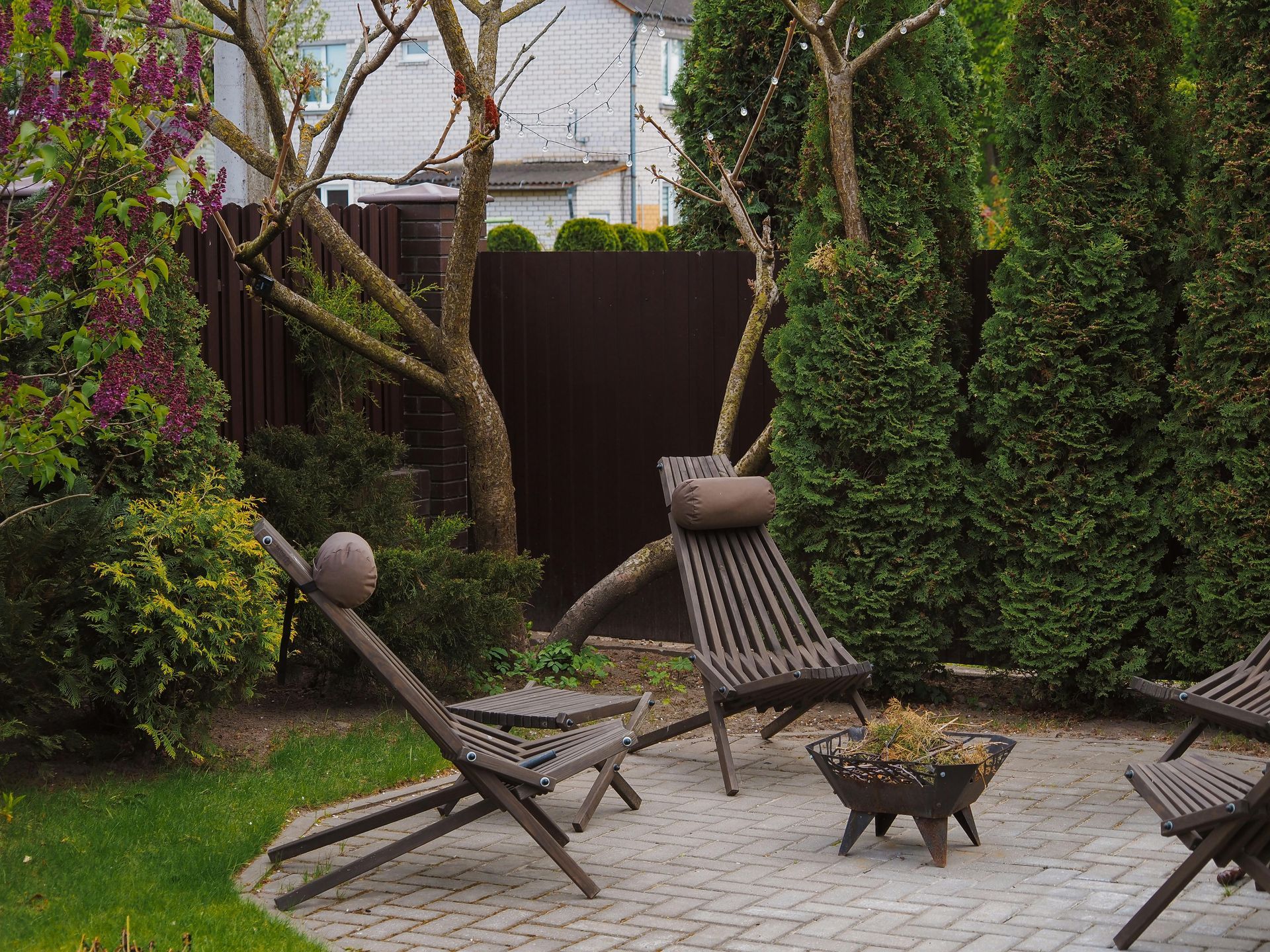 A small backyard patio with lounge chairs, a fire pit, and tall hedges surrounding the space.
