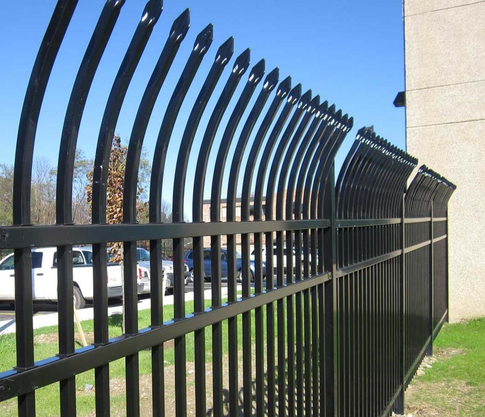 Black metal security fence with upward-curving posts and pointed finials, outdoors on a sunny day.
