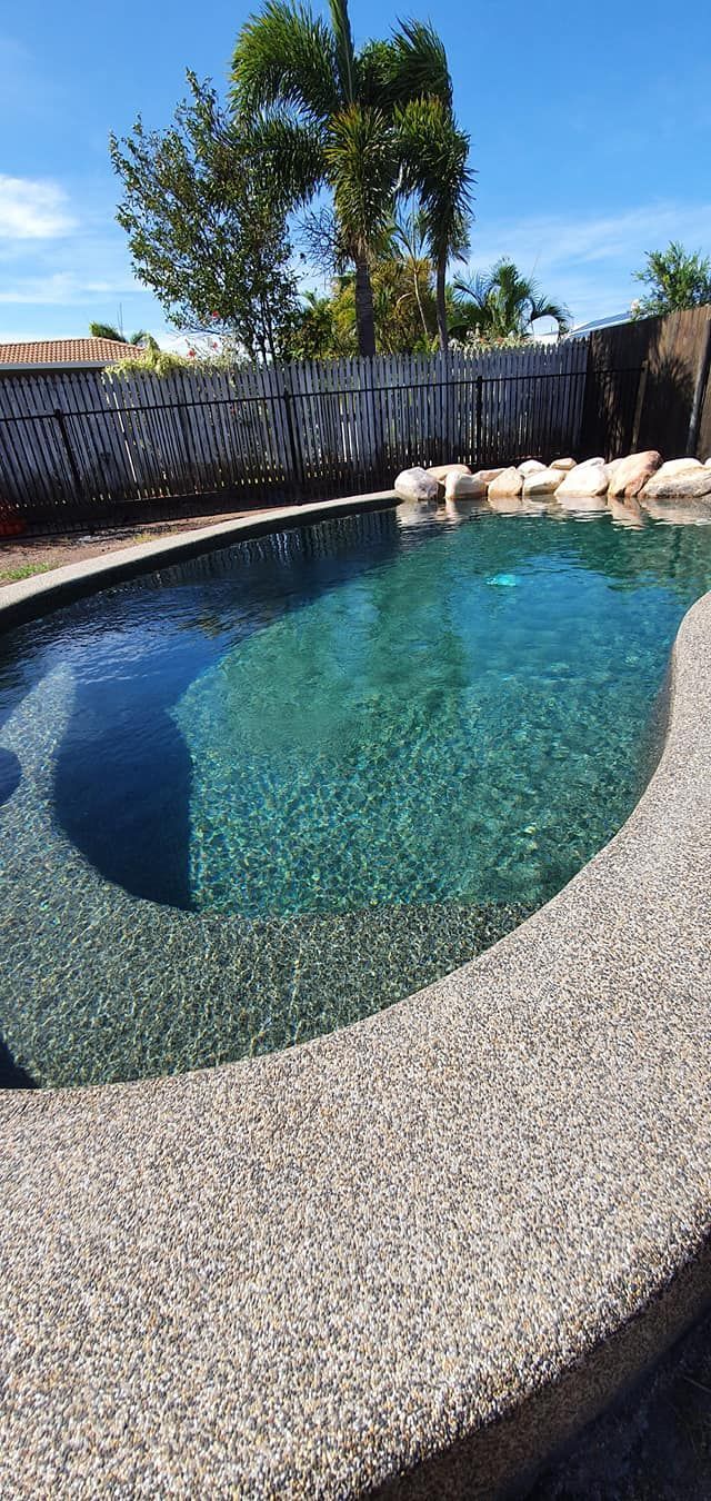A Large Swimming Pool Surrounded by Rocks and Trees on a Sunny Day — NQ Tropical Pools & Renovations in Burdekin, QLD