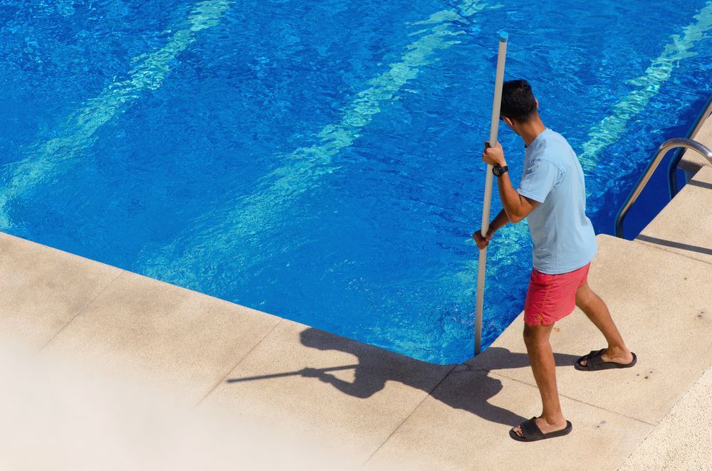 A Man is Cleaning a Swimming Pool With a Broom — NQ Tropical Pools & Renovations in Ingham, QLD