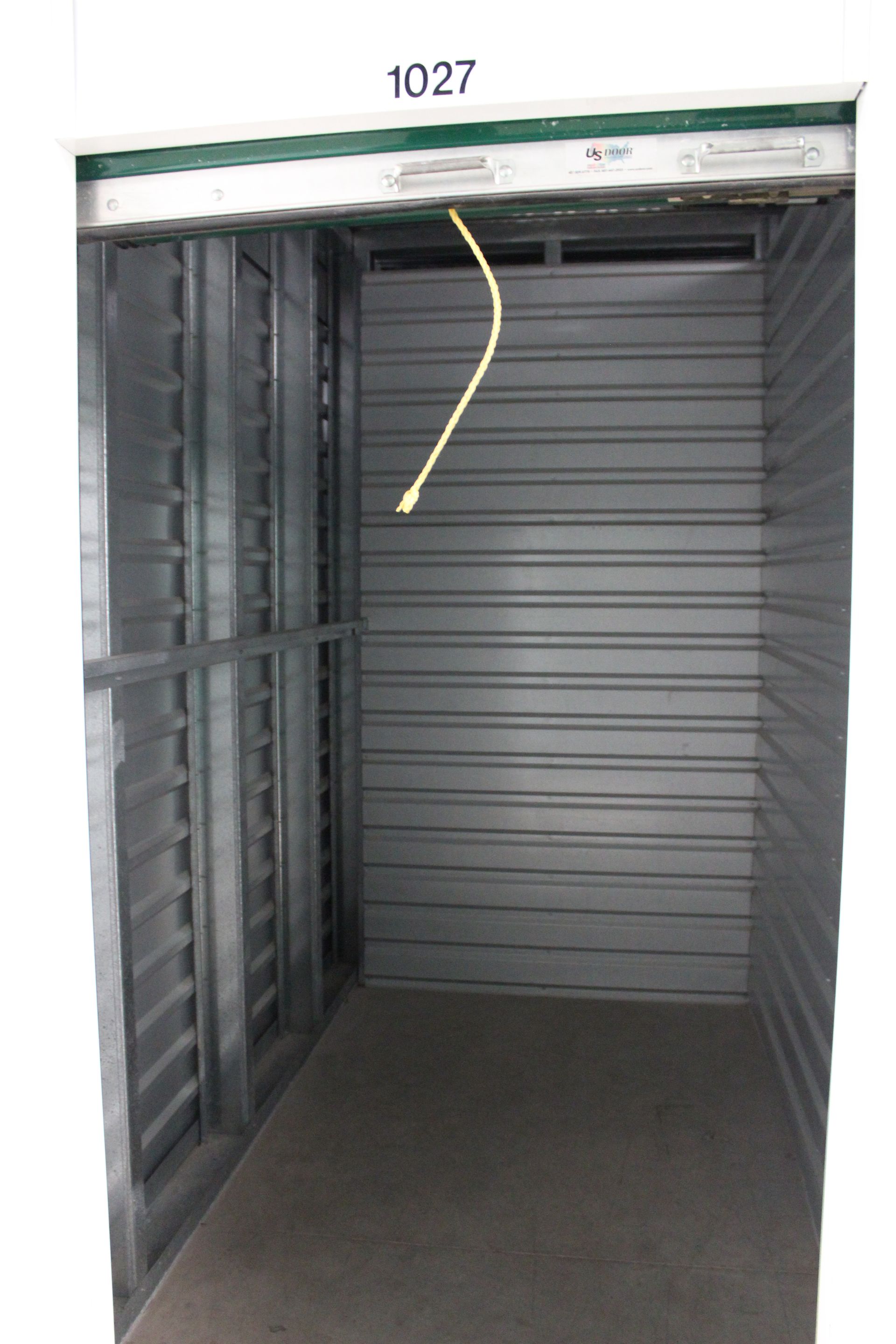 Empty, open storage unit, metallic walls and door, concrete floor. Unit number 1027 visible.