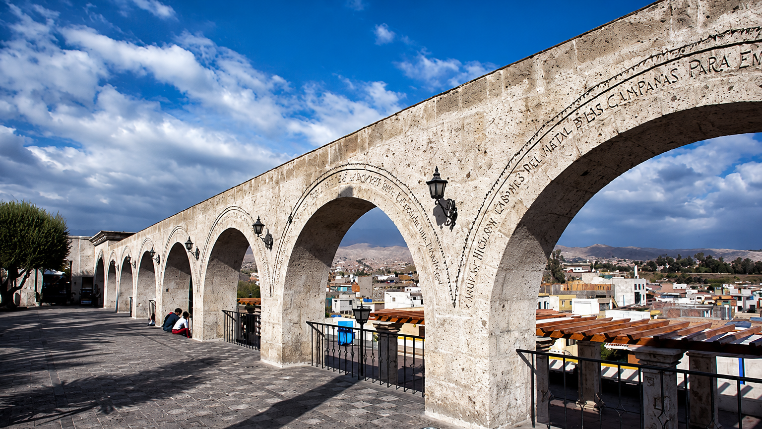 Acueducto de arco de piedra con vistas a un pueblo en la ladera de una colina bajo un cielo azul.