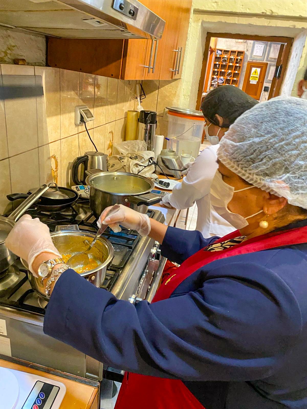 Dos personas con delantales y redecillas para el pelo cocinando en una estufa en una cocina, revolviendo ollas y sartenes.
