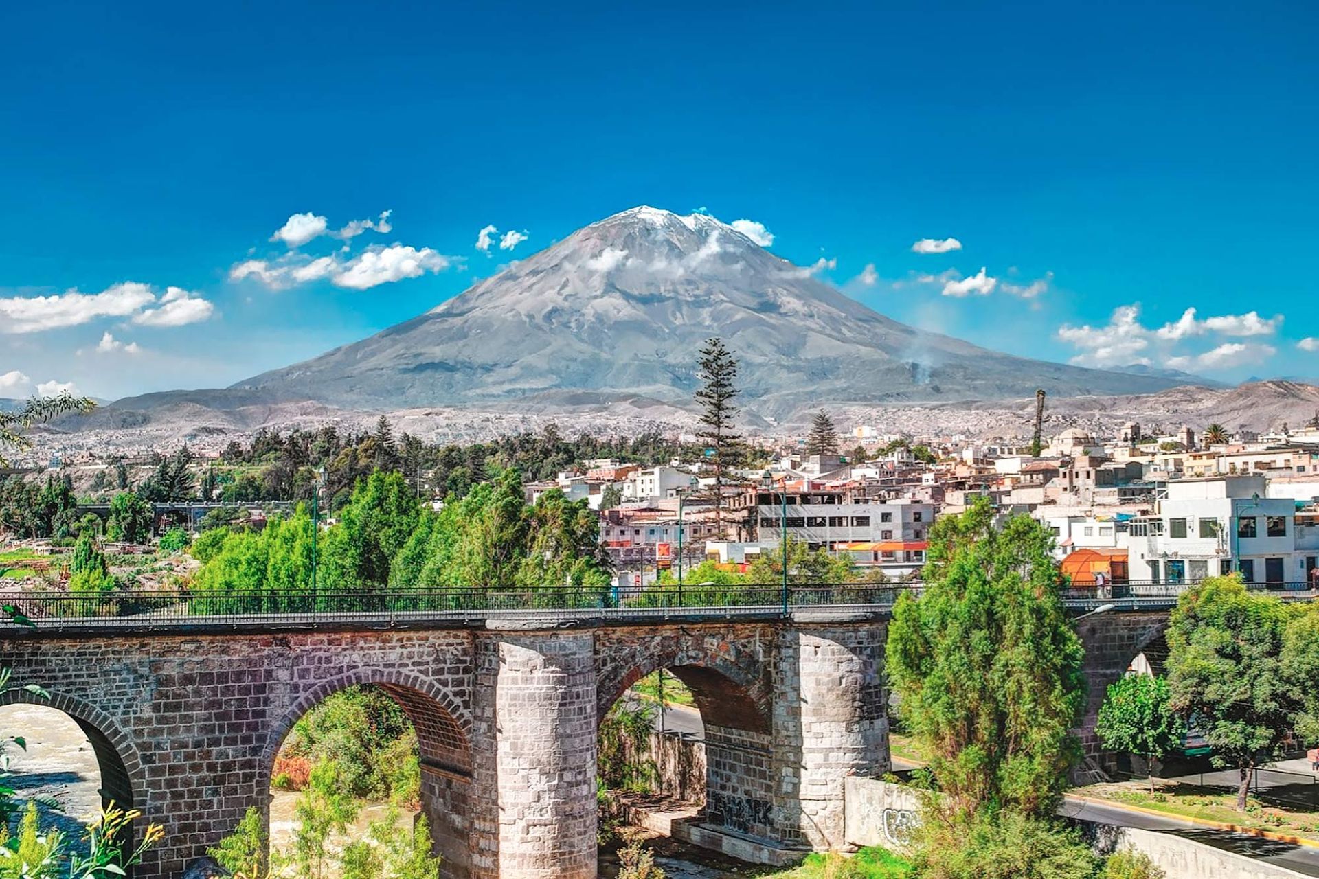 Puente de piedra sobre un valle verde con un pueblo y una montaña nevada bajo un cielo azul brillante.