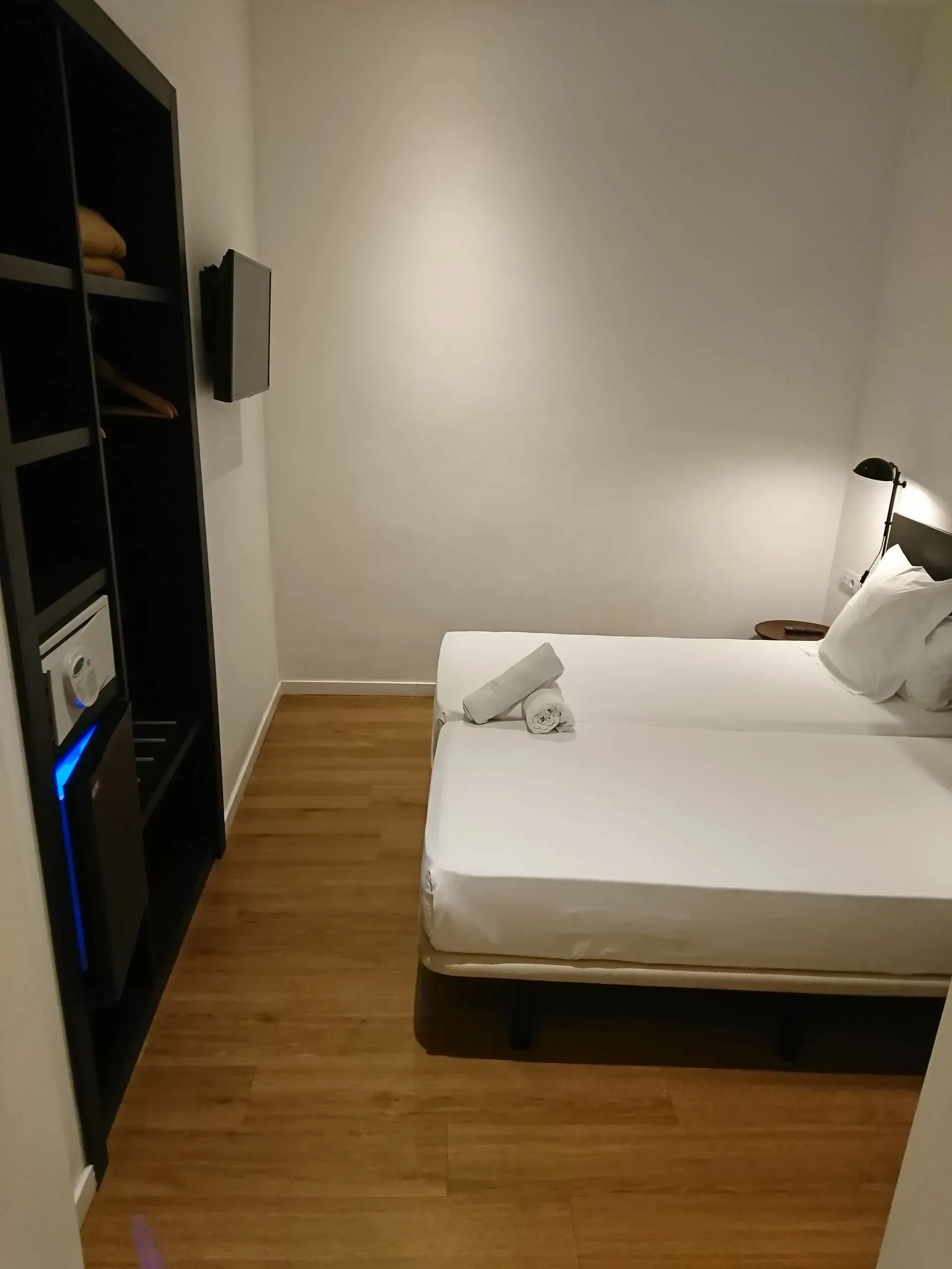 Una habitación de hotel con dos camas individuales blancas, una estantería negra con caja fuerte y un televisor en una pared.