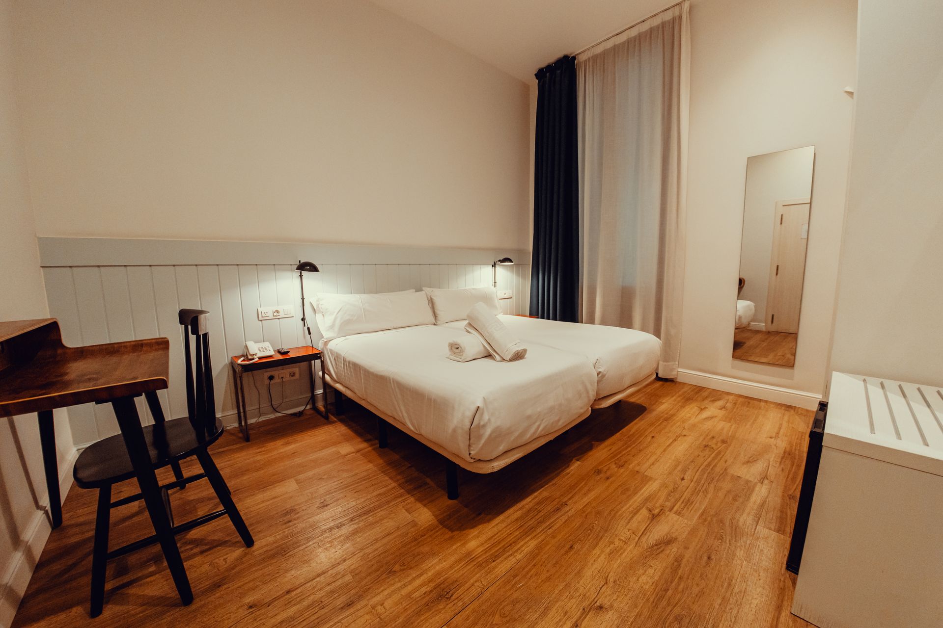 Habitación acogedora con cama doble, suelo de madera, lámparas de noche, escritorio, silla y espejo de pared.