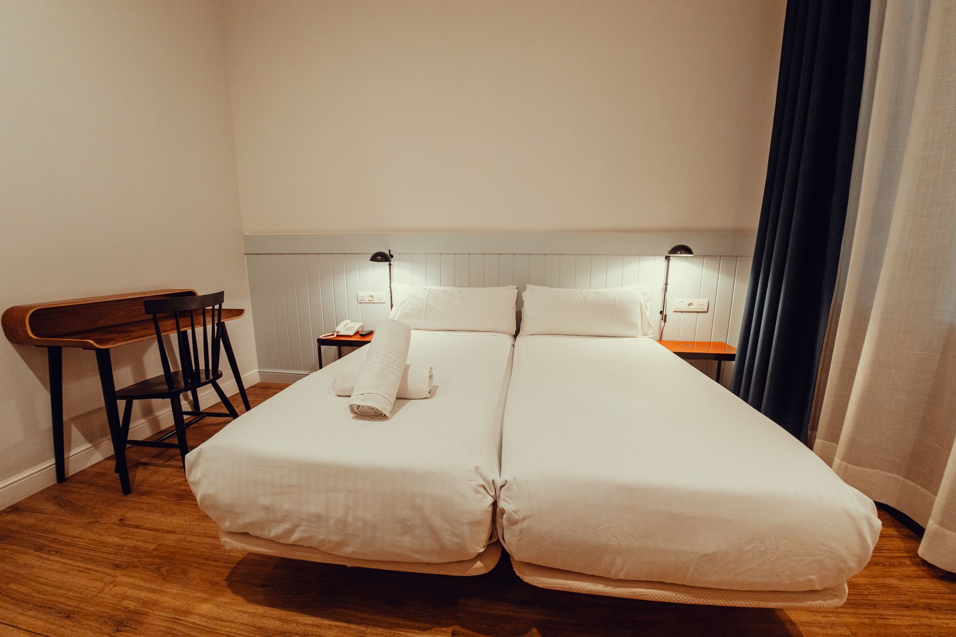 Habitación de hotel minimalista con dos camas blancas, lámparas de noche y un pequeño escritorio junto a la ventana.
