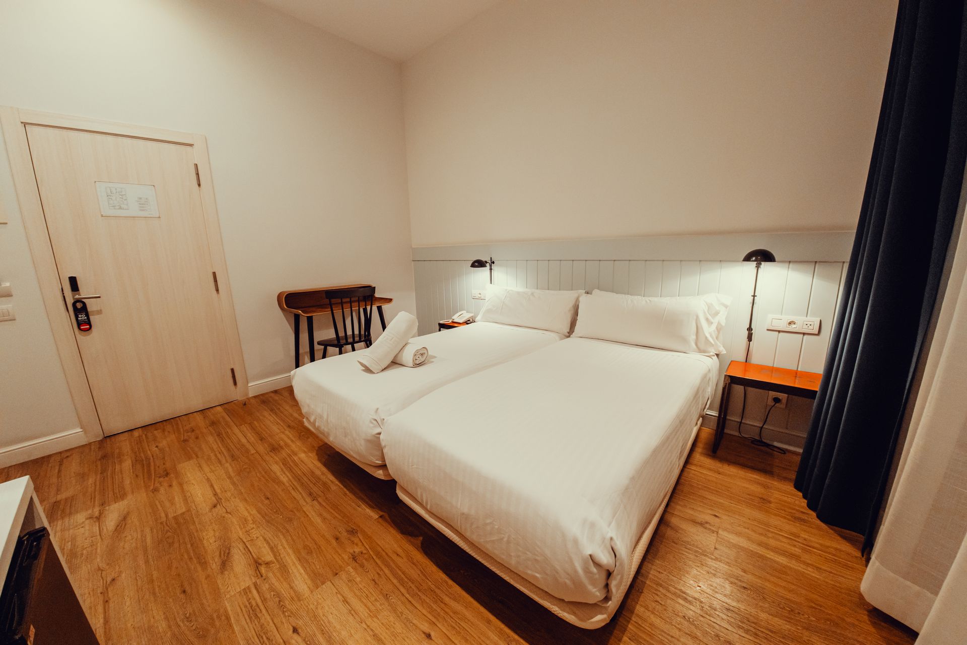 Dormitorio moderno con dos camas blancas, suelo de madera, lámparas de noche y una puerta cerrada.