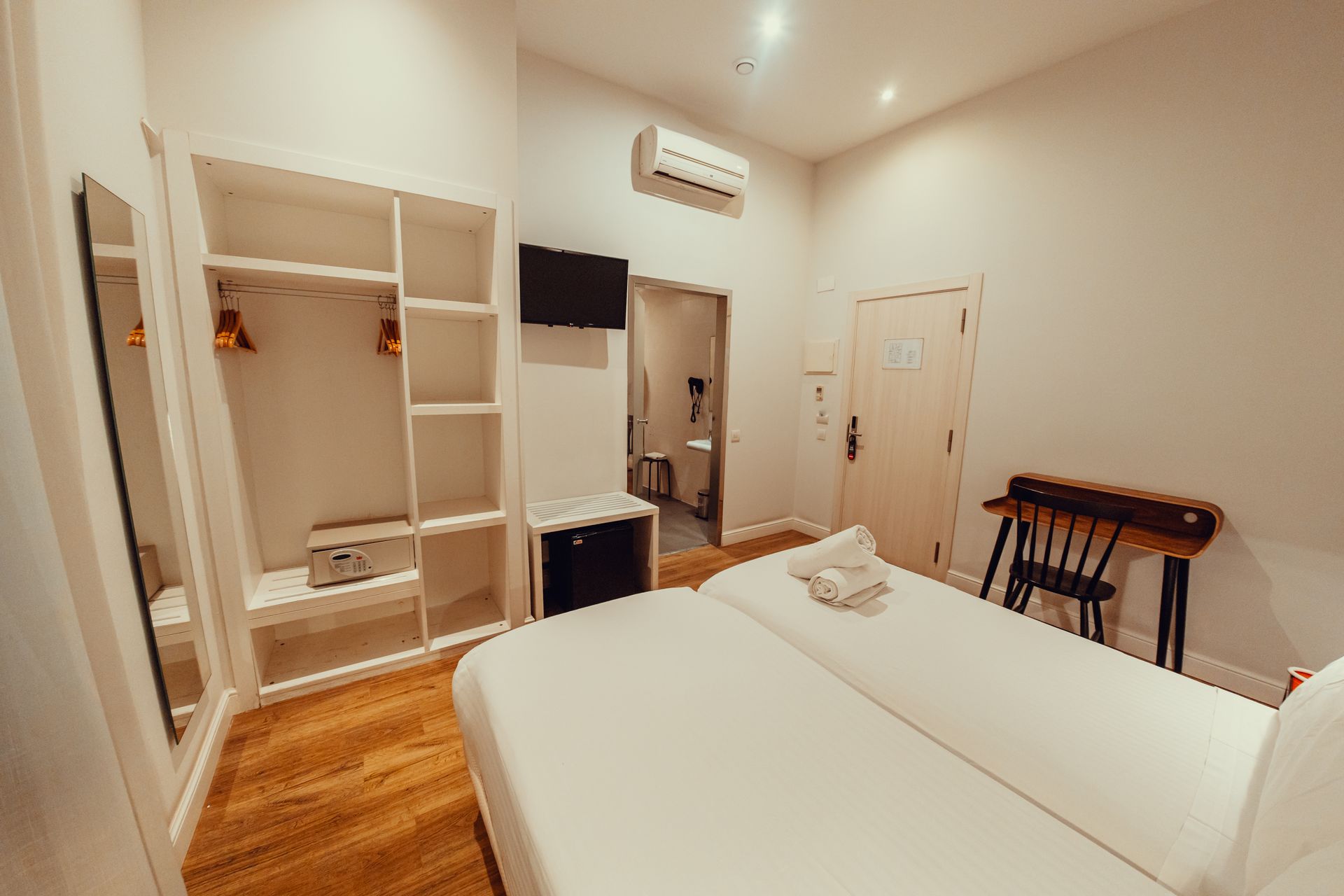 Habitación luminosa con dos camas, armario abierto, espejo de pared y mesita auxiliar bajo el aire acondicionado.