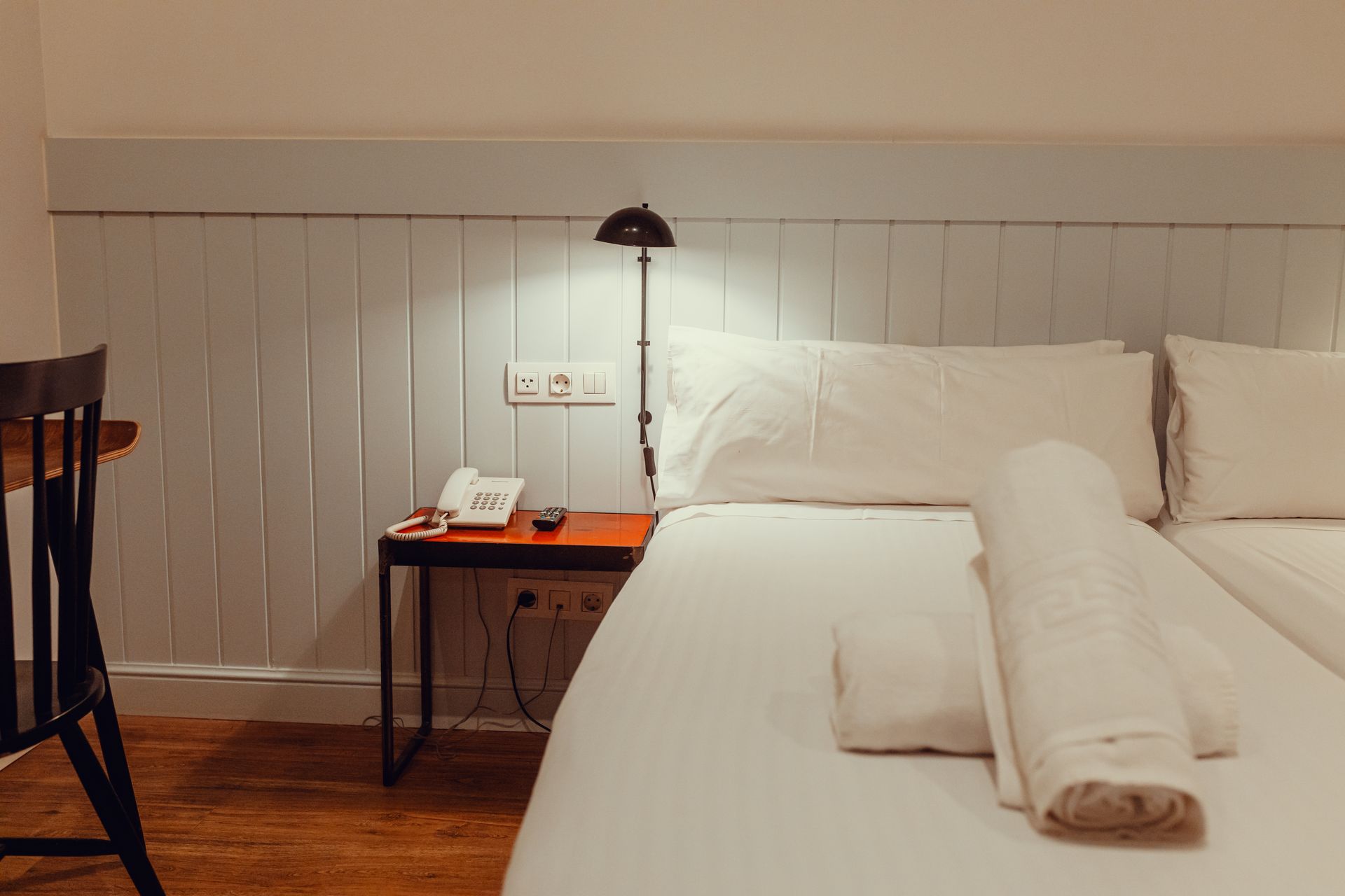Habitación acogedora con cama blanca, toallas dobladas, lámpara de noche y una pequeña mesita de noche de madera.