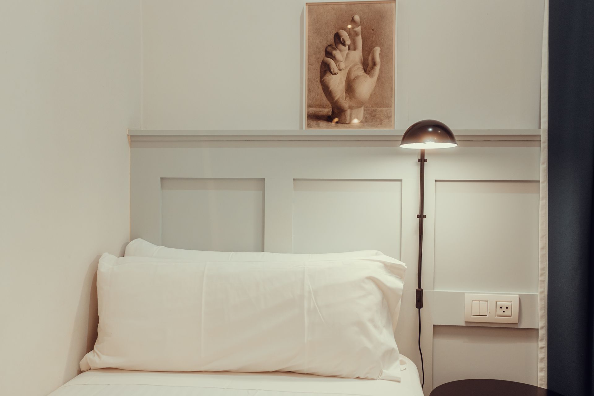Dormitorio minimalista con cama blanca, cabecero gris, lámpara de noche y cuadro enmarcado sobre la cama.