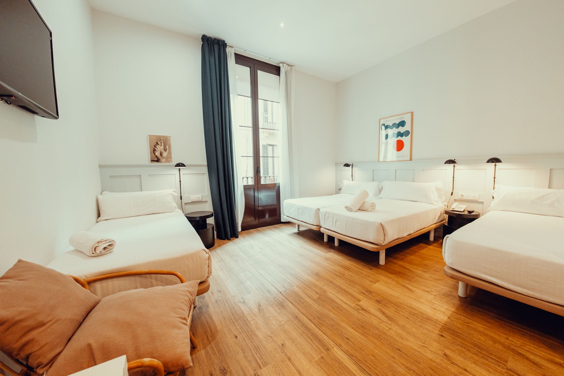 Habitación luminosa con tres camas, suelo de madera y ventana con cortinas oscuras.