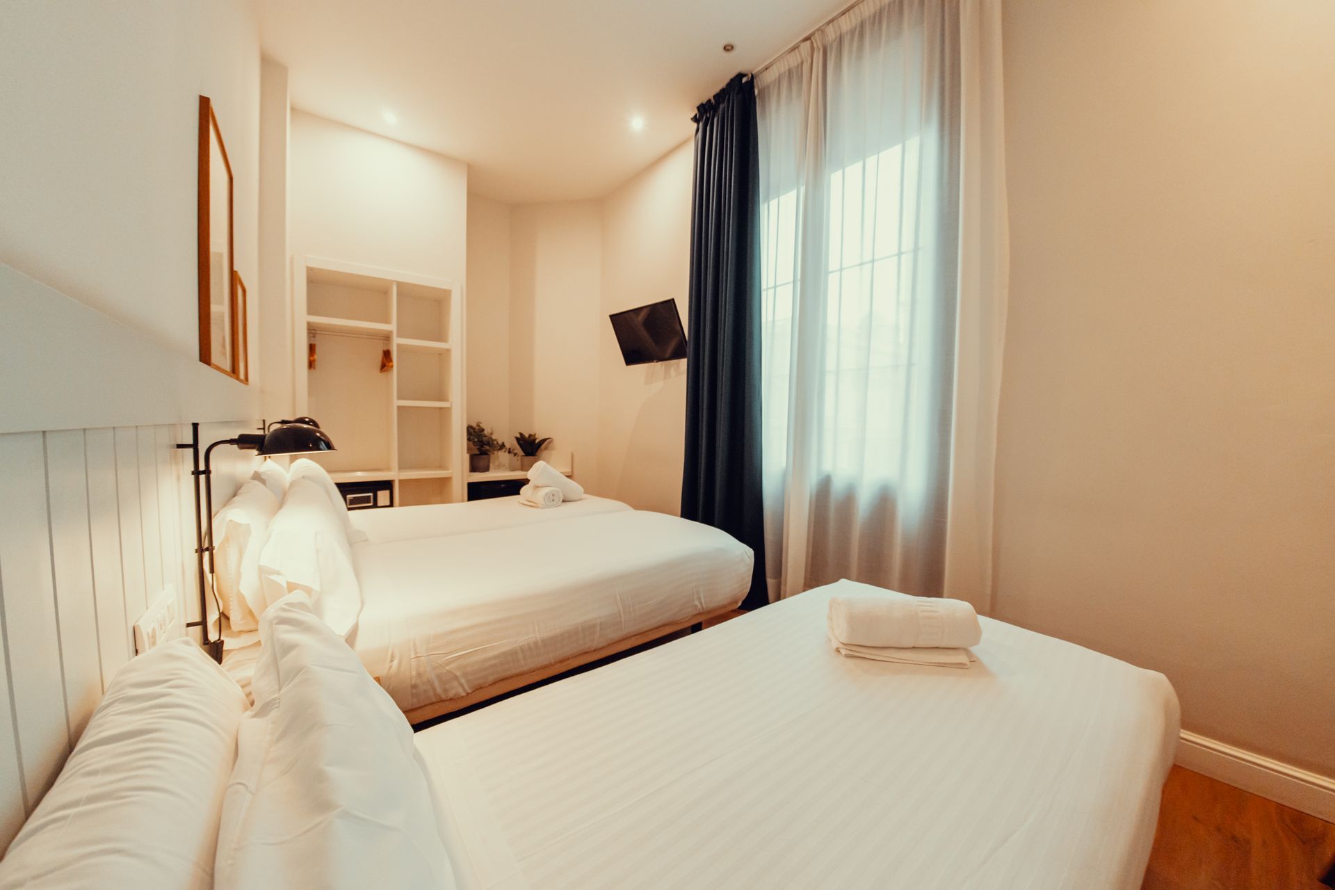 Habitación doble luminosa con dos camas, sábanas blancas y una gran ventana con cortinas oscuras.