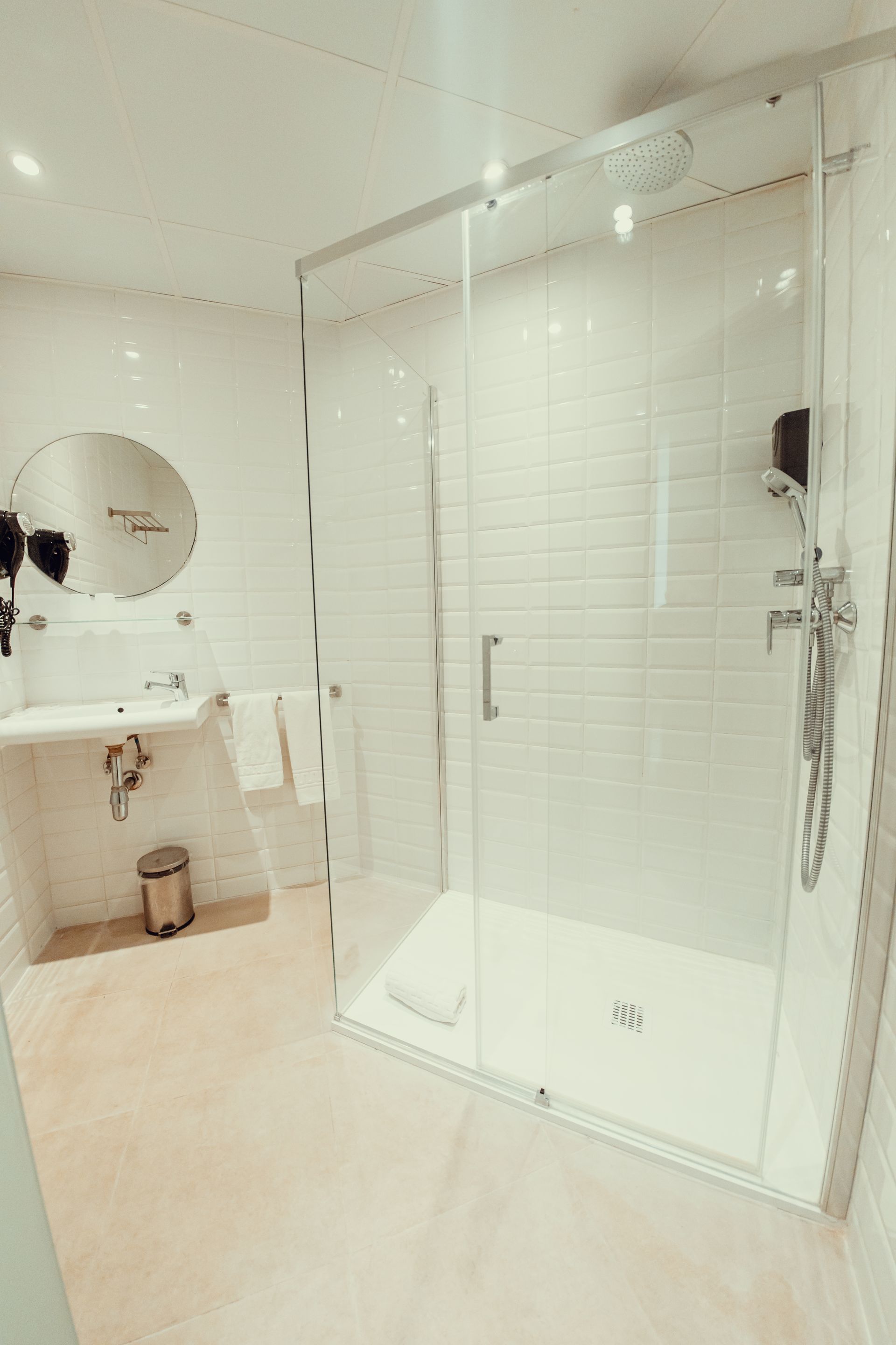 Baño luminoso con ducha a ras de suelo de cristal, azulejos blancos, lavabo y espejo redondo.