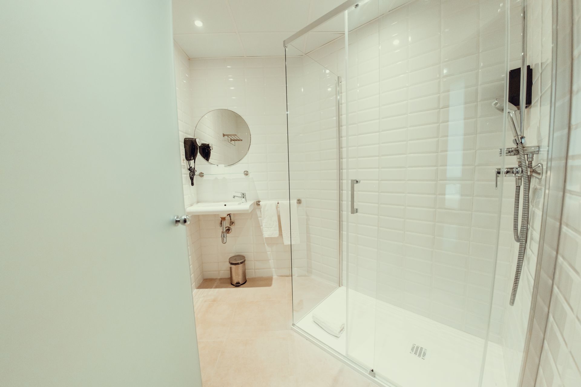 Baño moderno y luminoso con ducha de cristal, lavabo, espejo y suelo de baldosas beige.