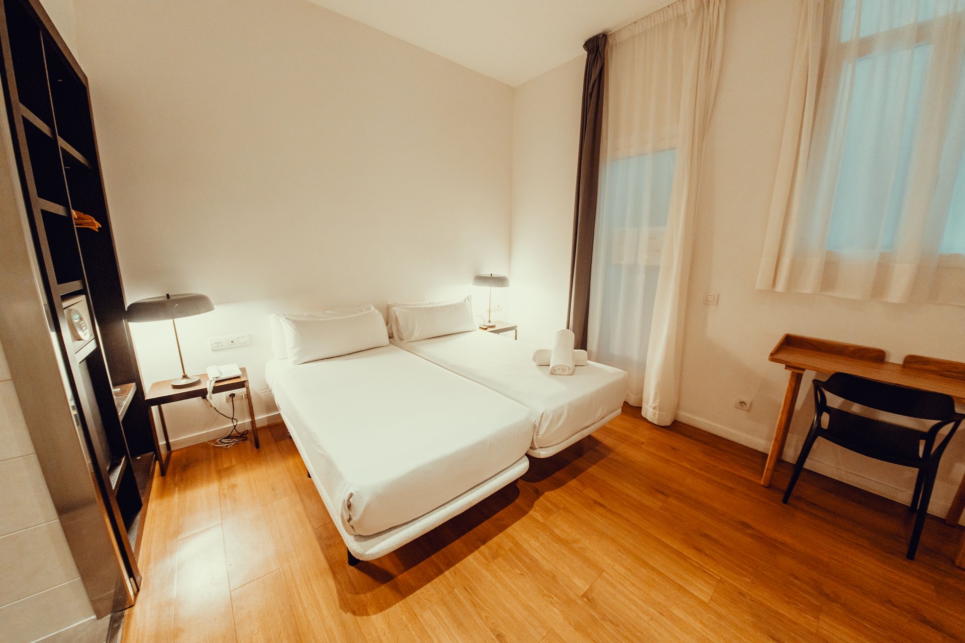 Habitación luminosa con cama blanca, suelo de madera, escritorio y puertas de balcón con cortinas.