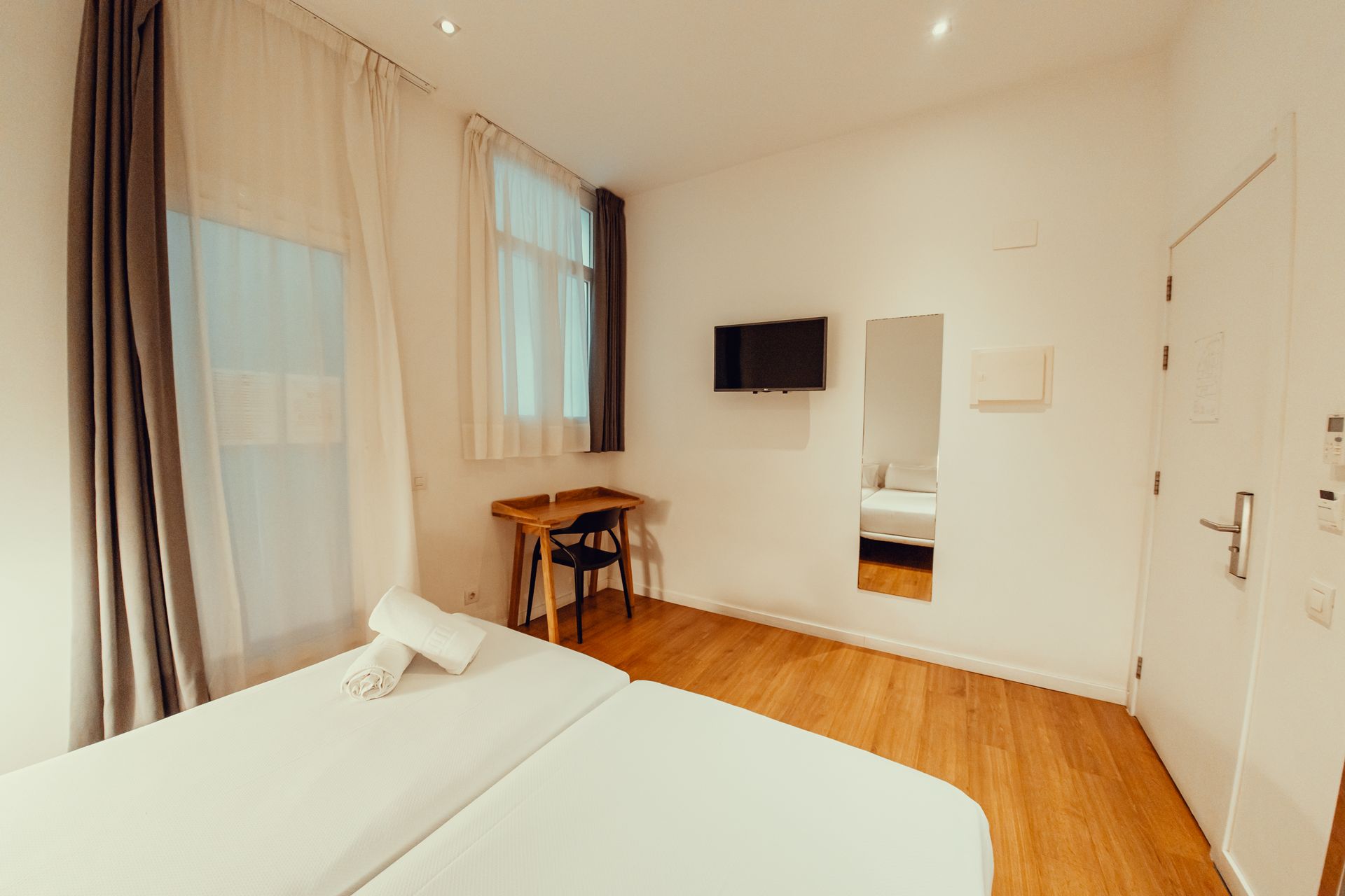 Habitación luminosa y minimalista con cama, televisor de pared, escritorio, espejo y grandes ventanas con cortinas.